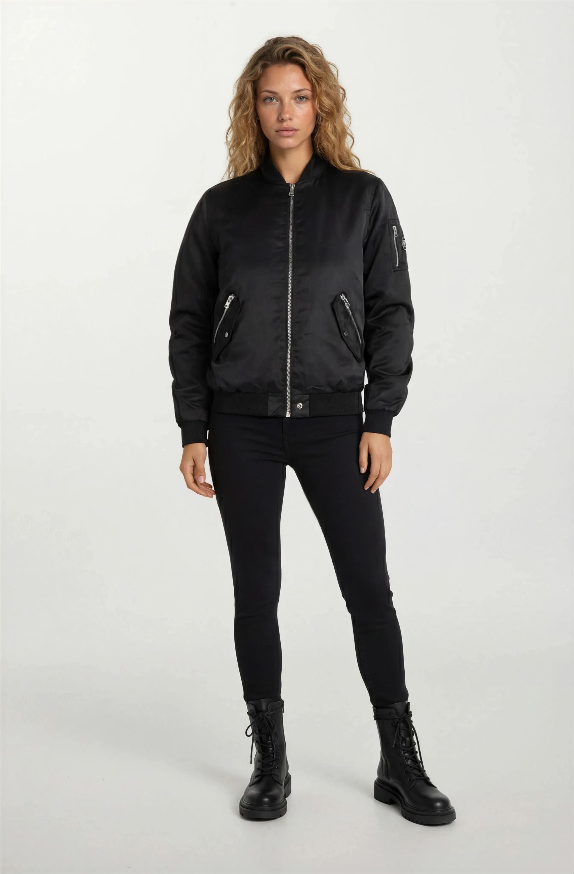 Eine Frau mit langen gewellten Haaren steht mit dem Gesicht nach vorne und trägt eine trueprodigy Elegante Damen Bomber Jacke mit glänzender Oberfläche, eine schwarze Hose und Schnürstiefel vor einem weißen Hintergrund. Diese Jacke verleiht ihrem ganz in Schwarz gehaltenen Look ein besonderes Flair.