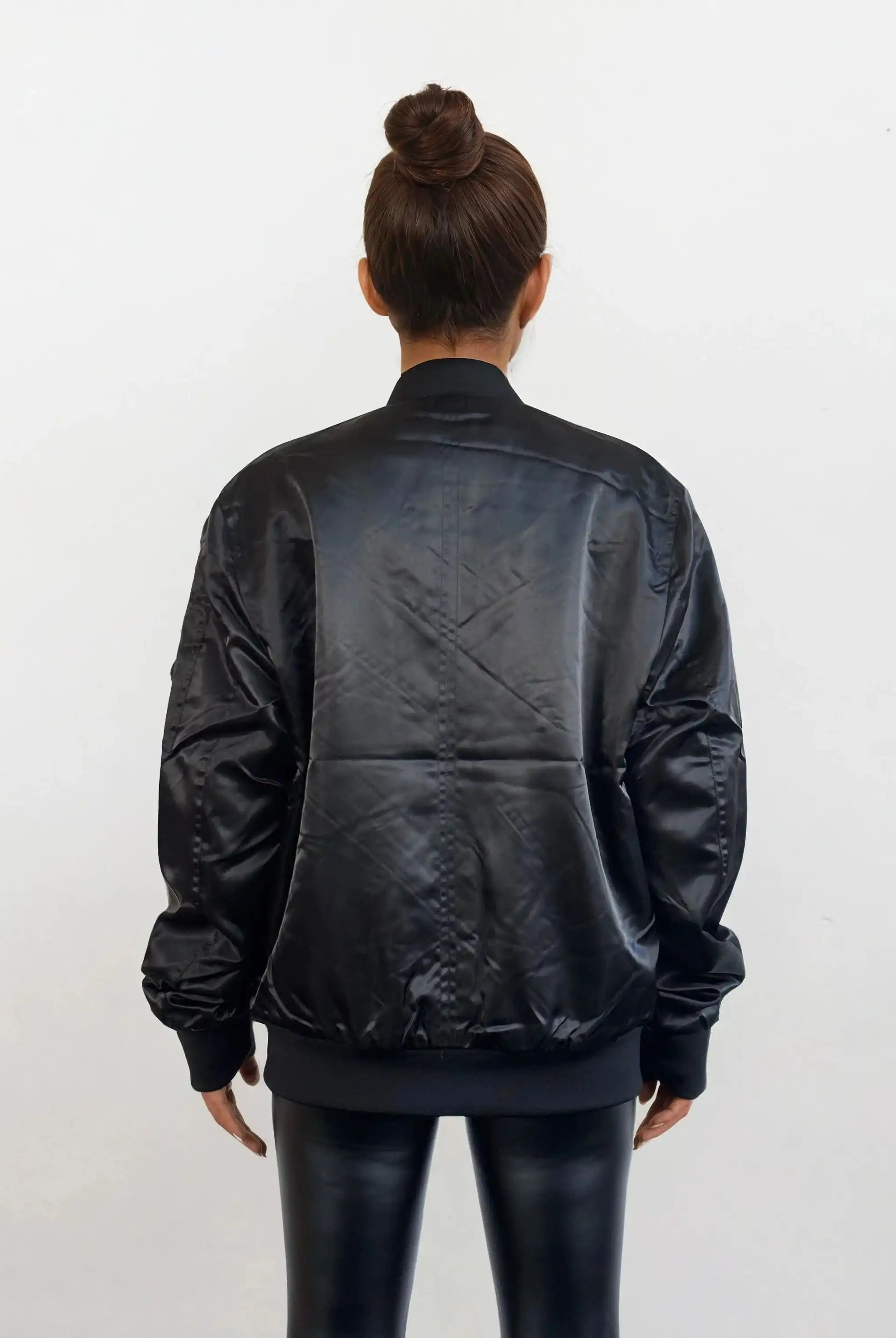 Eine Frau mit gepflegtem Dutt, von hinten gesehen vor einem weißen Hintergrund, trägt glänzende schwarze Leggings und die Elegante Damen Bomber Jacke mit glänzender Oberfläche von trueprodigy.