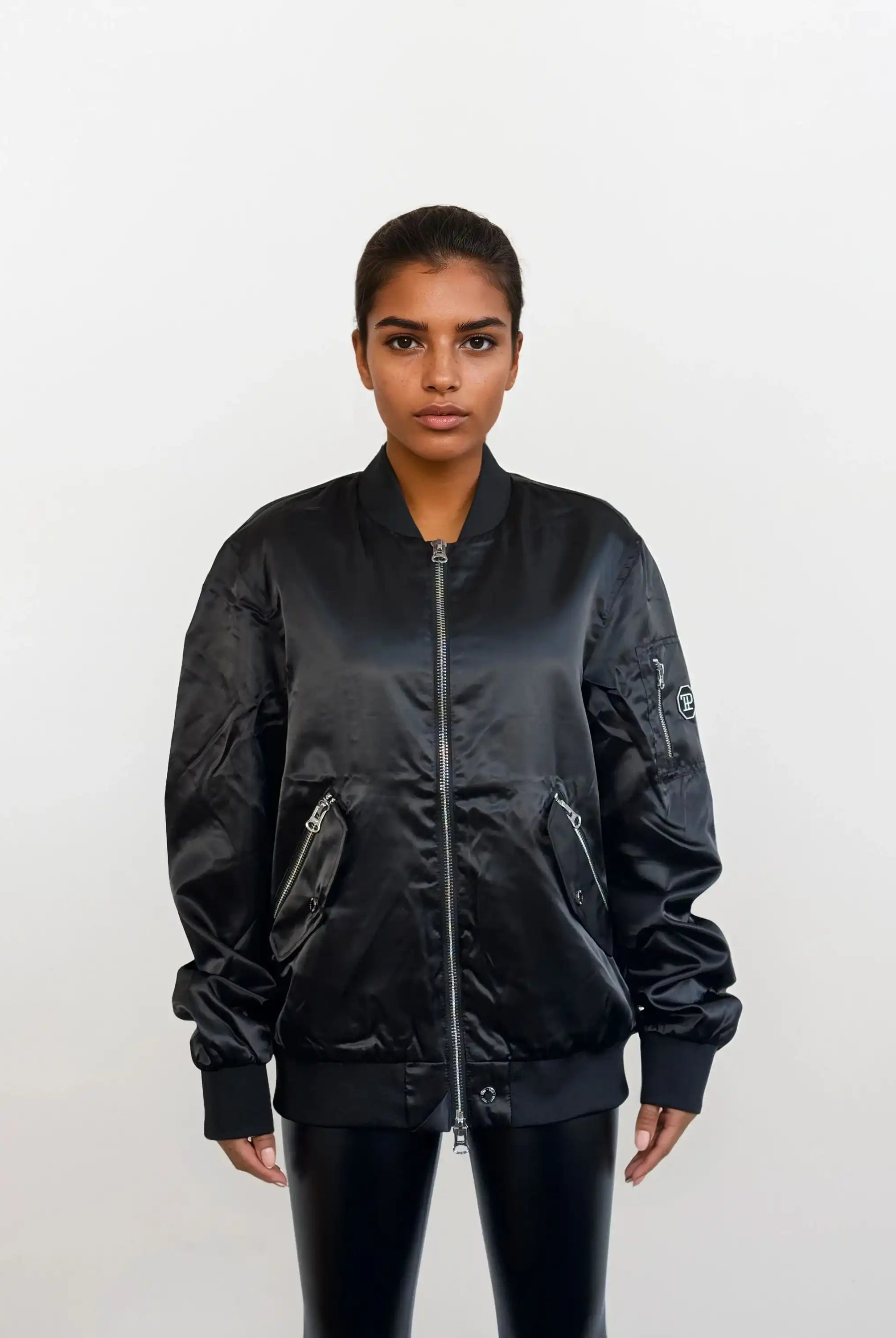 Eine Frau steht mit dem Gesicht nach vorne vor einem schlichten weißen Hintergrund und trägt eine trueprodigy Elegante Damen Bomberjacke mit glänzender Oberfläche, gepaart mit einer glänzenden schwarzen Hose.