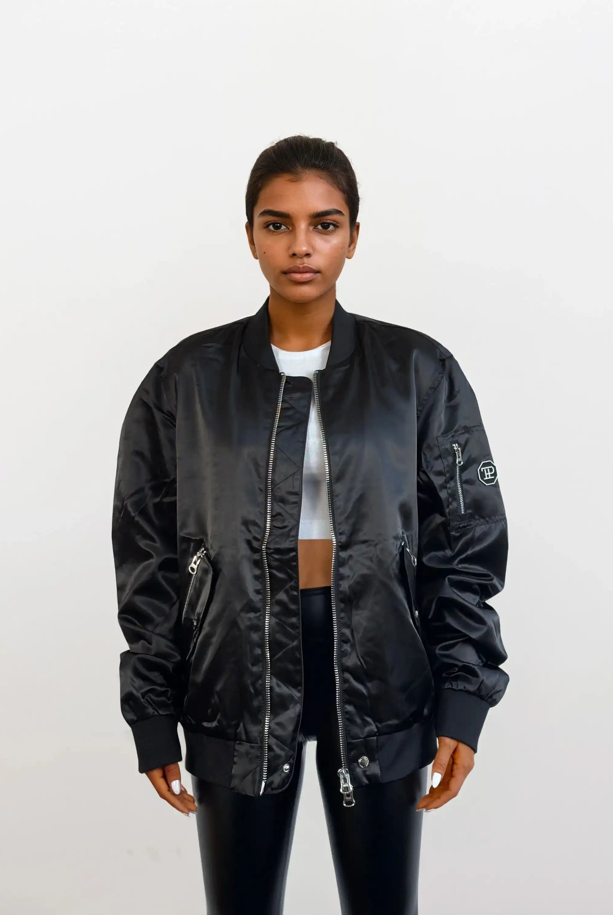 Eine Person steht mit dem Gesicht nach vorne vor einem einfarbig weißen Hintergrund und trägt eine trueprodigy Elegante Damen Bomberjacke mit glänzender Oberfläche über einem weißen, kurz geschnittenen Oberteil und einer schwarzen Hose. Ihr Haar ist zurückgesteckt, und ihr Gesichtsausdruck ist neutral.