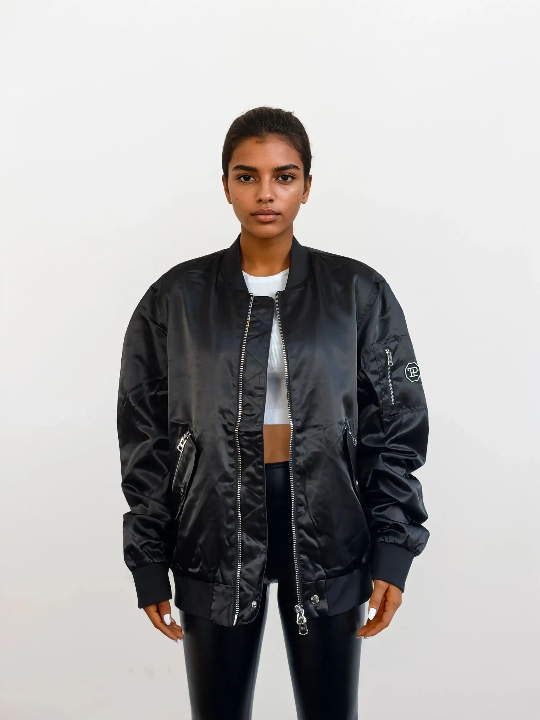 Eine Person steht mit dem Gesicht nach vorne vor einem einfarbig weißen Hintergrund und trägt eine trueprodigy Elegante Damen Bomberjacke mit glänzender Oberfläche über einem weißen, kurz geschnittenen Oberteil und einer schwarzen Hose. Ihr Haar ist zurückgesteckt, und ihr Gesichtsausdruck ist neutral.