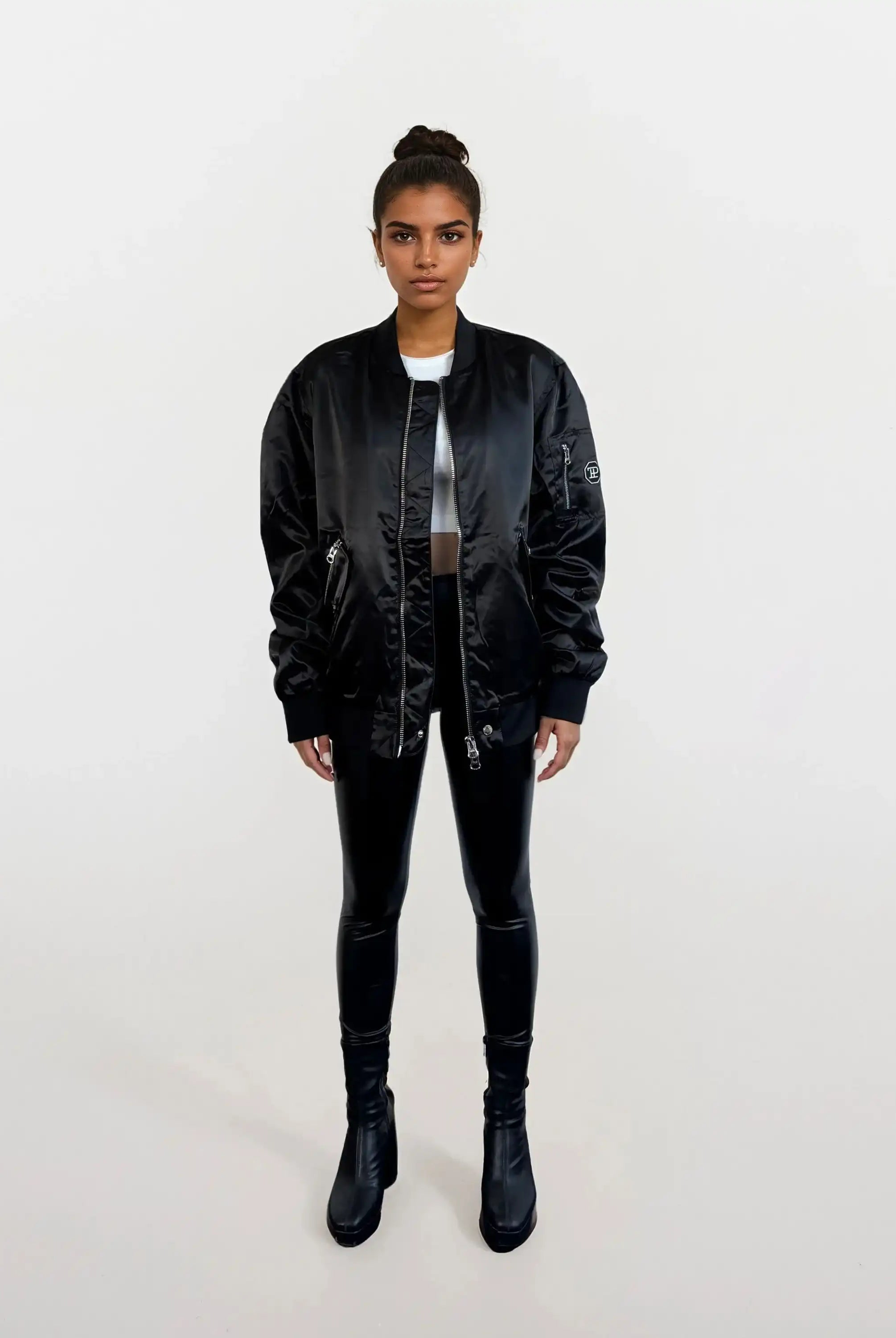 Eine Person steht mit dem Gesicht nach vorne vor einem weißen Hintergrund, trägt eine schwarze trueprodigy Elegante Damen Bomberjacke mit glänzender Oberfläche, schwarz glänzende Leggings, ein weißes Oberteil, schwarze Stiefel und hat die Haare zu einem Dutt gebunden.