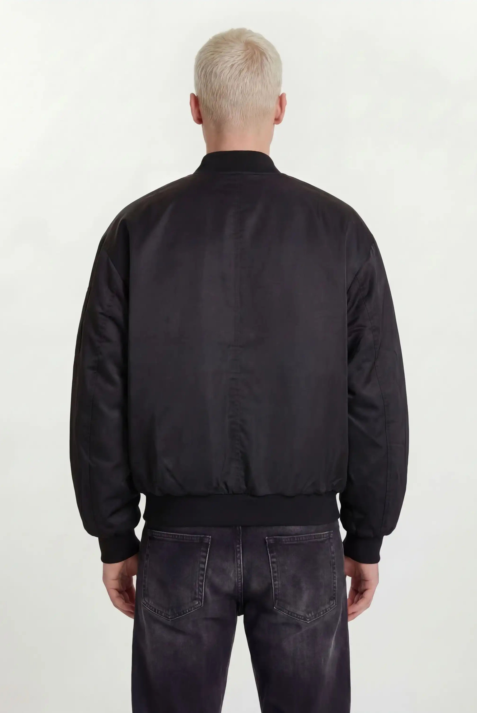 Eine Person mit kurzen, hellblonden Haaren ist von hinten zu sehen. Sie trägt eine trueprodigy Klassische Herren Bomberjacke mit glänzender Oberfläche und dunkel gewaschene Jeans, die vor einem schlichten, hellen Hintergrund für einen urbanen Style-Look stehen.