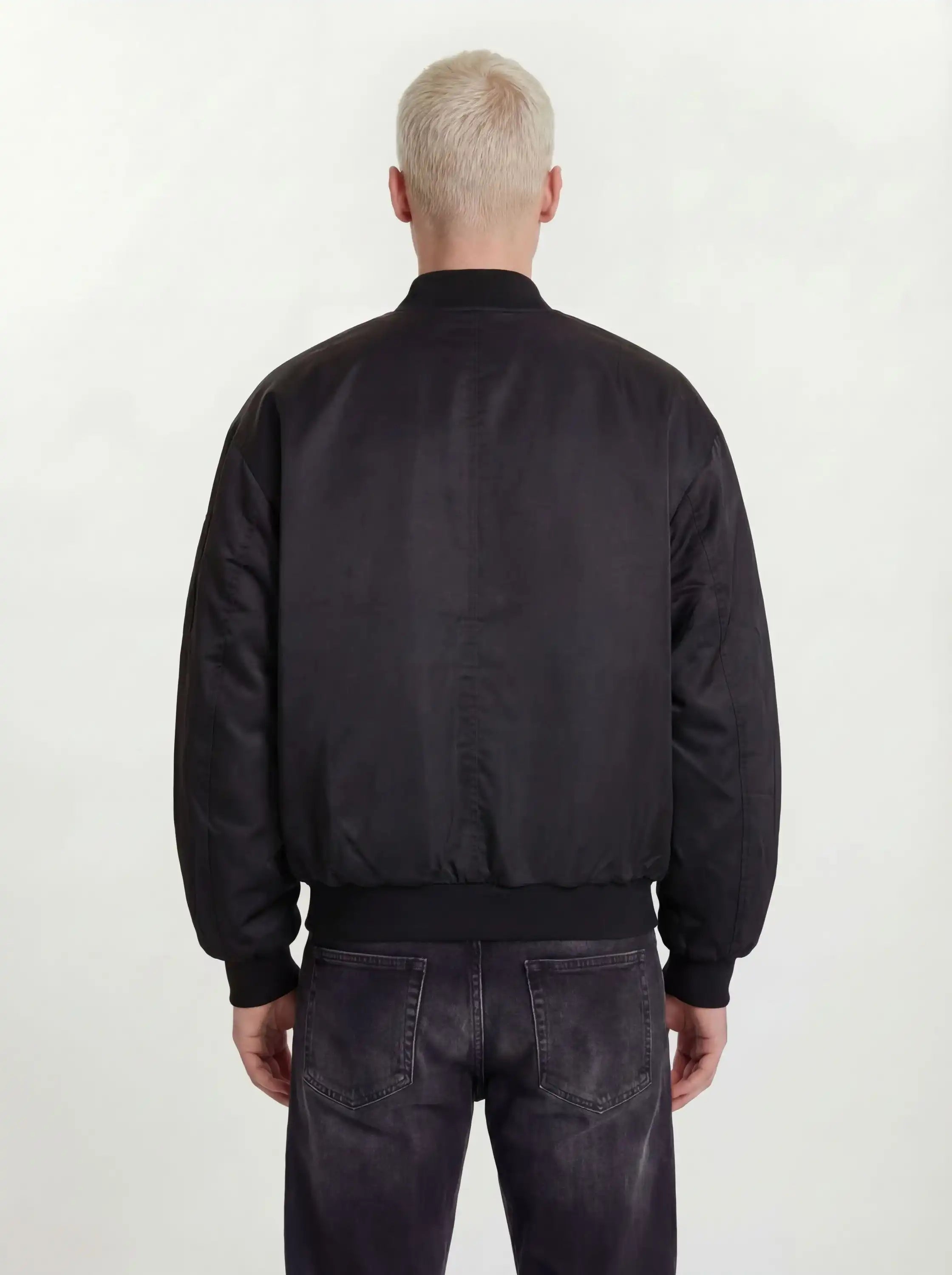 Eine Person mit kurzen, hellblonden Haaren ist von hinten zu sehen. Sie trägt eine trueprodigy Klassische Herren Bomberjacke mit glänzender Oberfläche und dunkel gewaschene Jeans, die vor einem schlichten, hellen Hintergrund für einen urbanen Style-Look stehen.