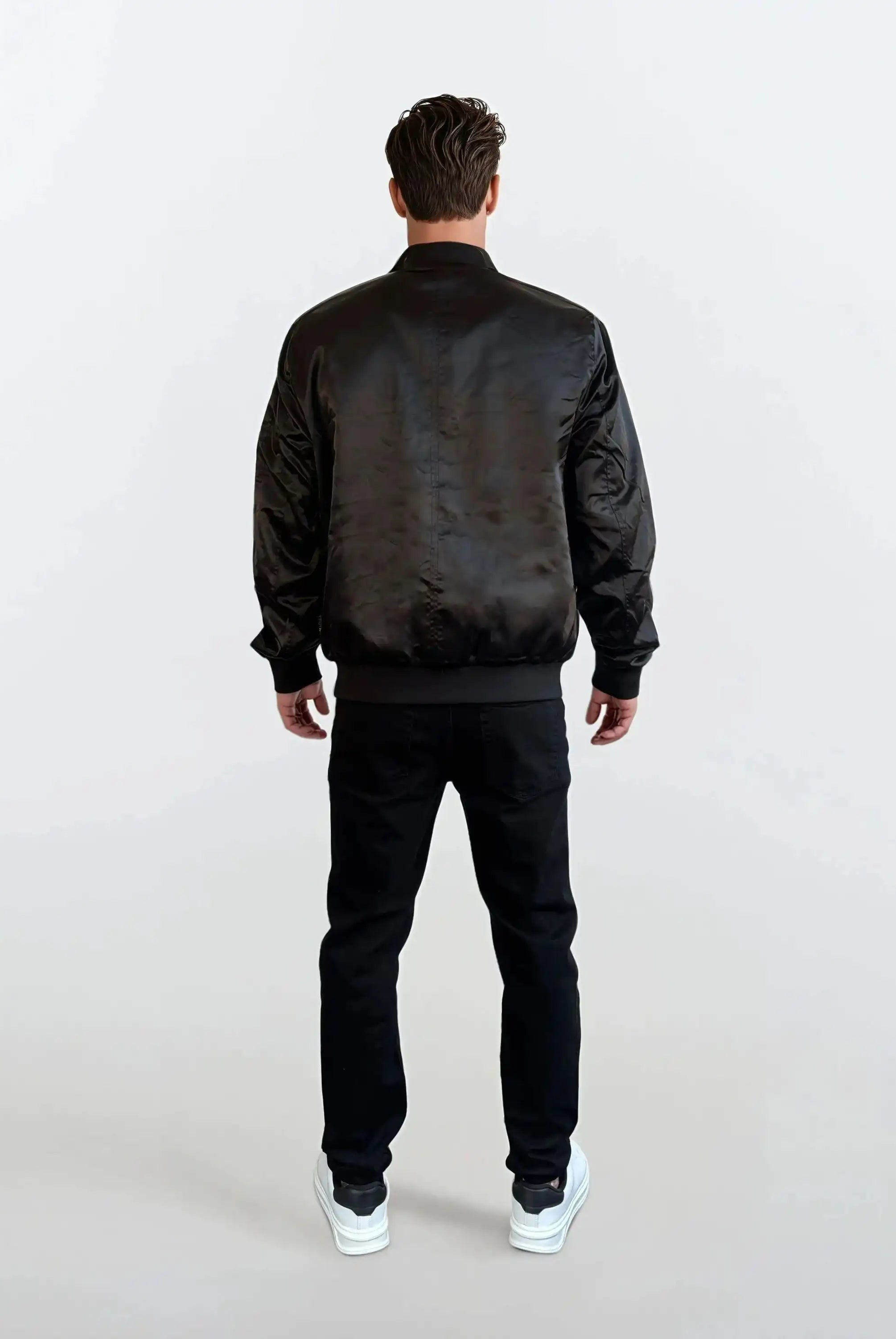 Eine Person steht mit dem Rücken zur Kamera und trägt die Klassische Herren Bomber Jacke mit glänzender Oberfläche von trueprodigy, schwarzen Hosen und weißen Sneakers - ein urbaner Look vor hellem, schlichten Hintergrund.