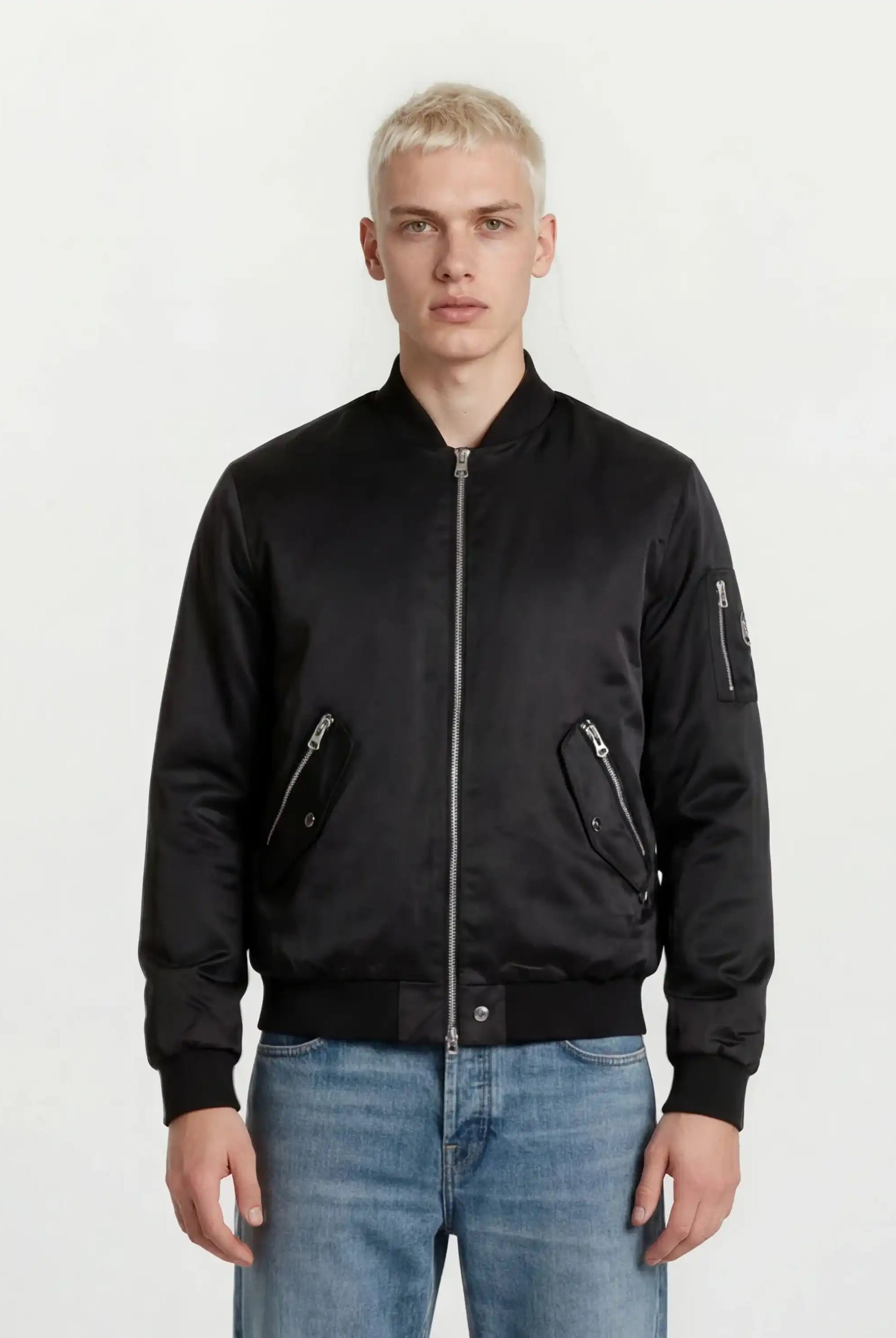 Eine Person mit kurzen blonden Haaren modelliert urbaner Style in einer trueprodigy Klassische Herren Bomber Jacke mit glänzender Oberfläche und hellblauen Jeans, stehend vor einem schlichten weißen Hintergrund.