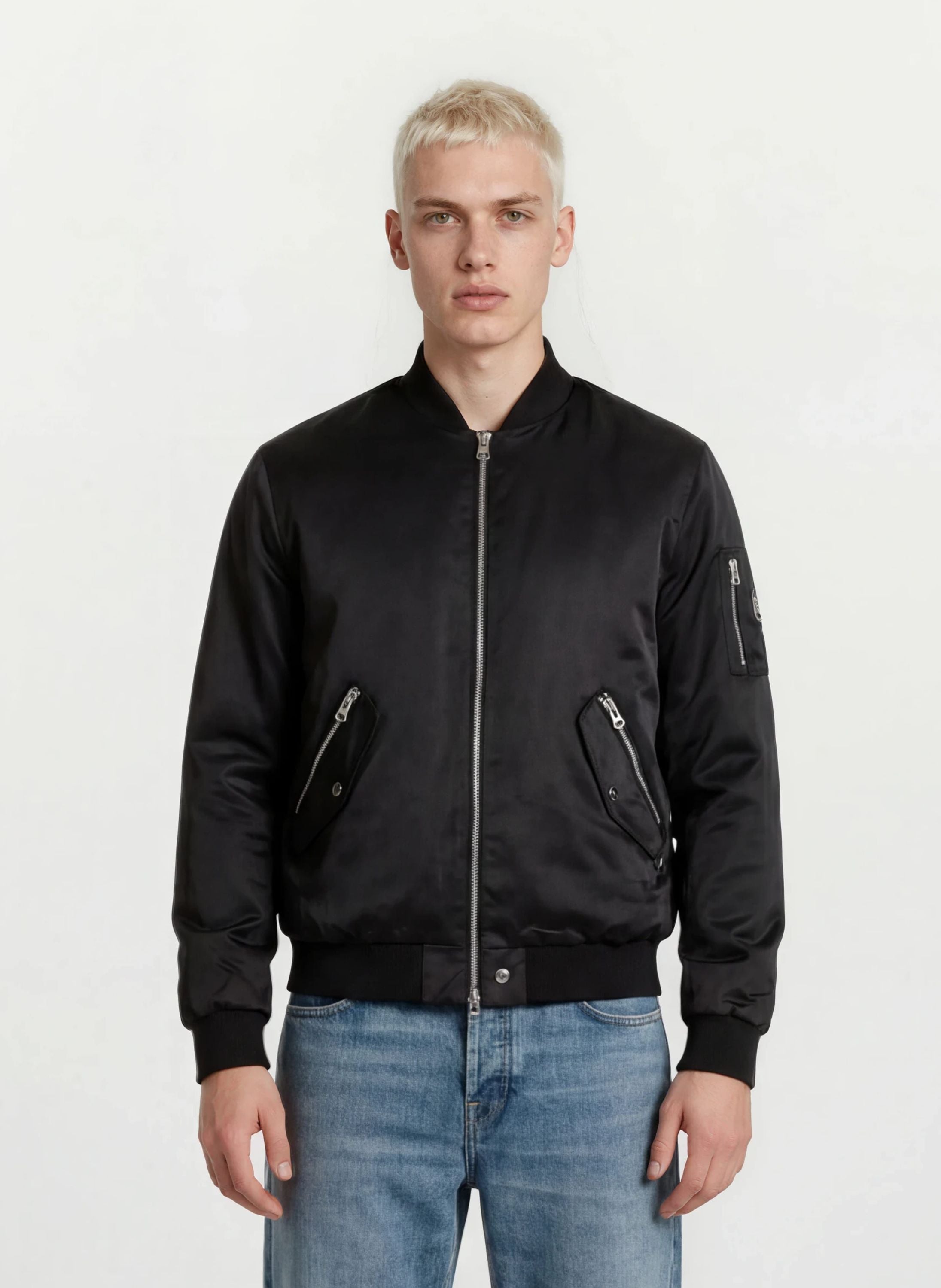 Eine Person mit kurzen blonden Haaren modelliert urbaner Style in einer trueprodigy Klassische Herren Bomber Jacke mit glänzender Oberfläche und hellblauen Jeans, stehend vor einem schlichten weißen Hintergrund.