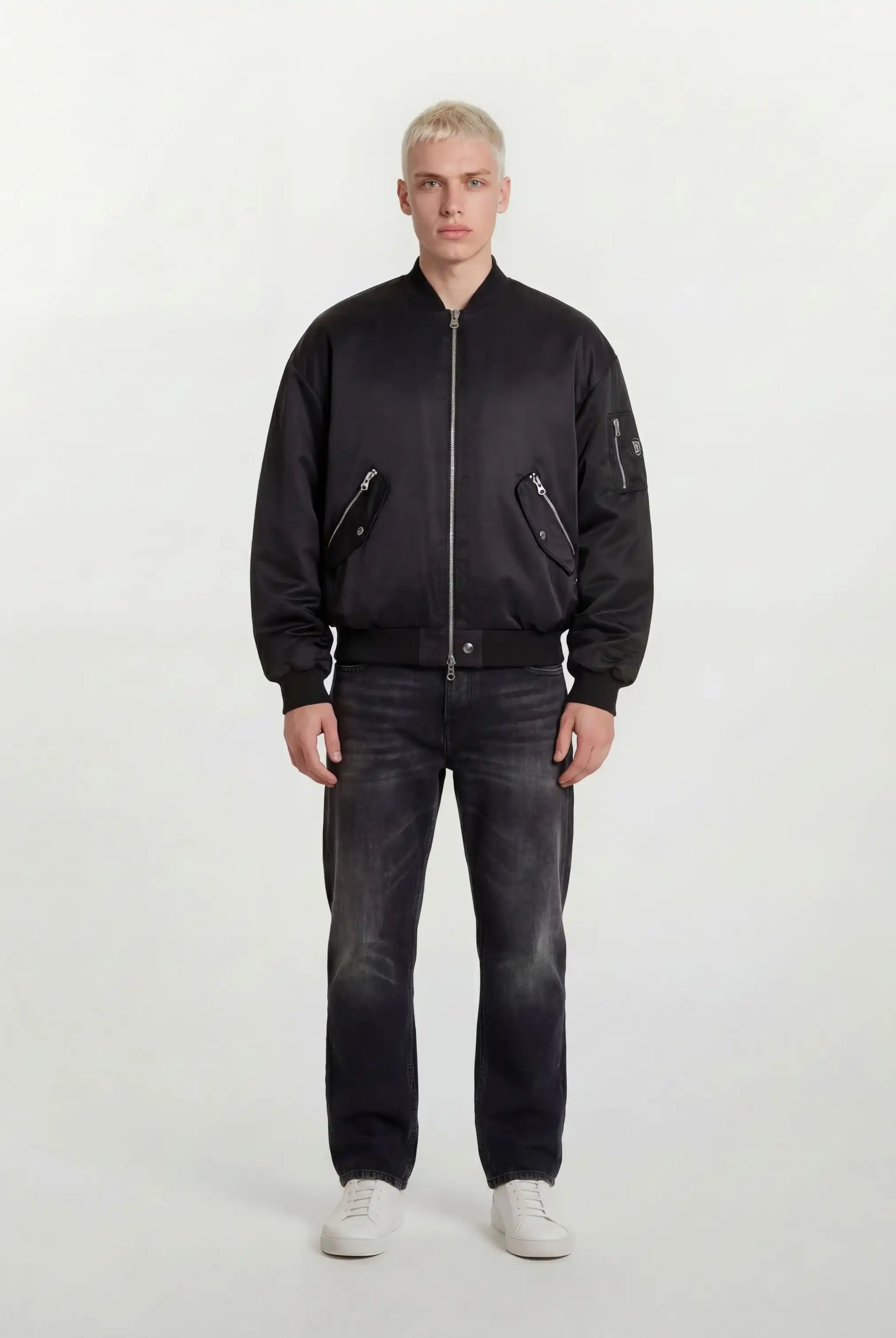 Eine Person mit kurzen blonden Haaren trägt eine trueprodigy Klassische Herren Bomberjacke mit glänzender Oberfläche, dunkle Jeans und weiße Turnschuhe und steht mit dem Gesicht nach vorne vor einem schlichten hellen Hintergrund.