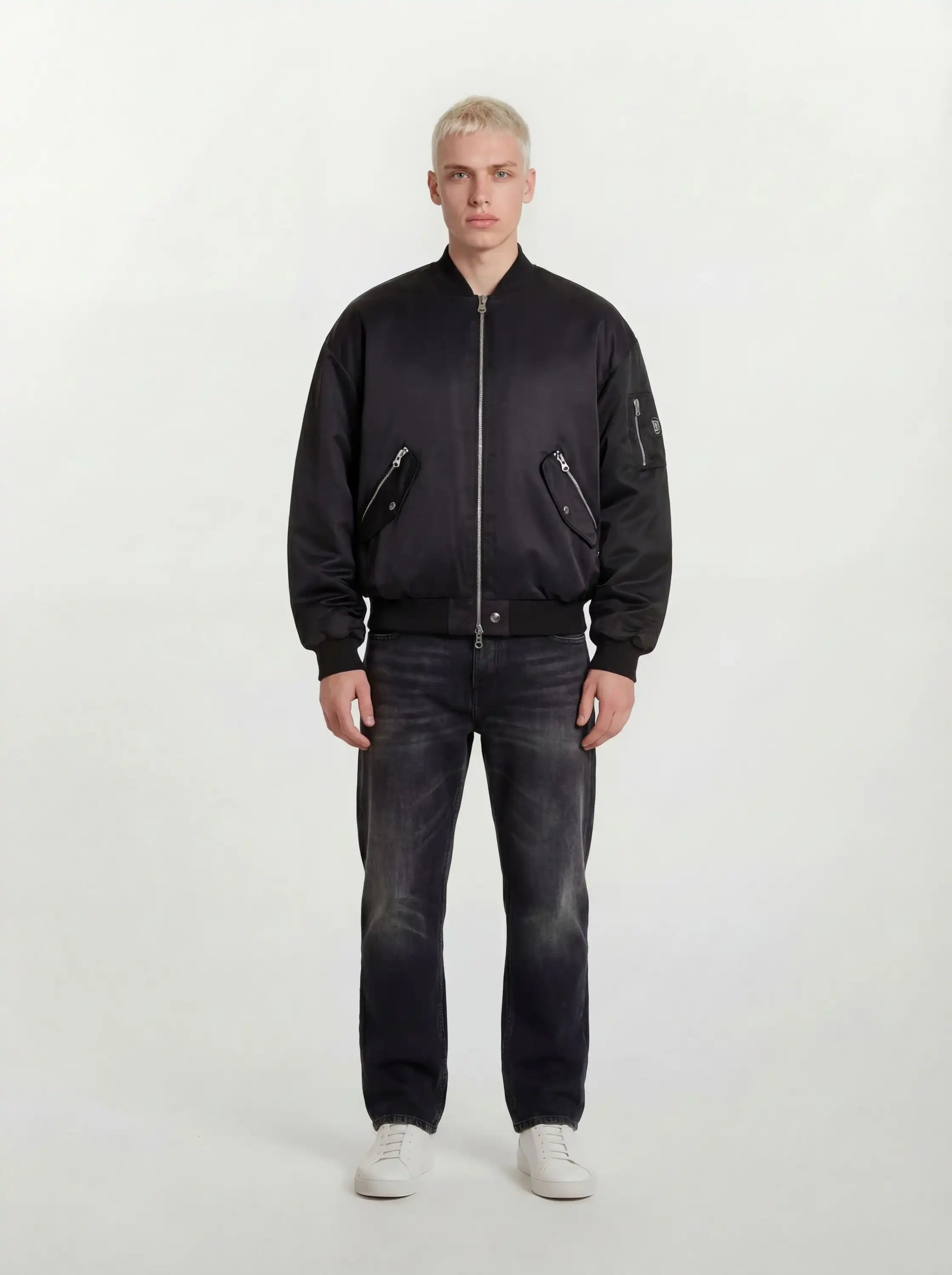 Eine Person mit kurzen blonden Haaren trägt eine trueprodigy Klassische Herren Bomberjacke mit glänzender Oberfläche, dunkle Jeans und weiße Turnschuhe und steht mit dem Gesicht nach vorne vor einem schlichten hellen Hintergrund.