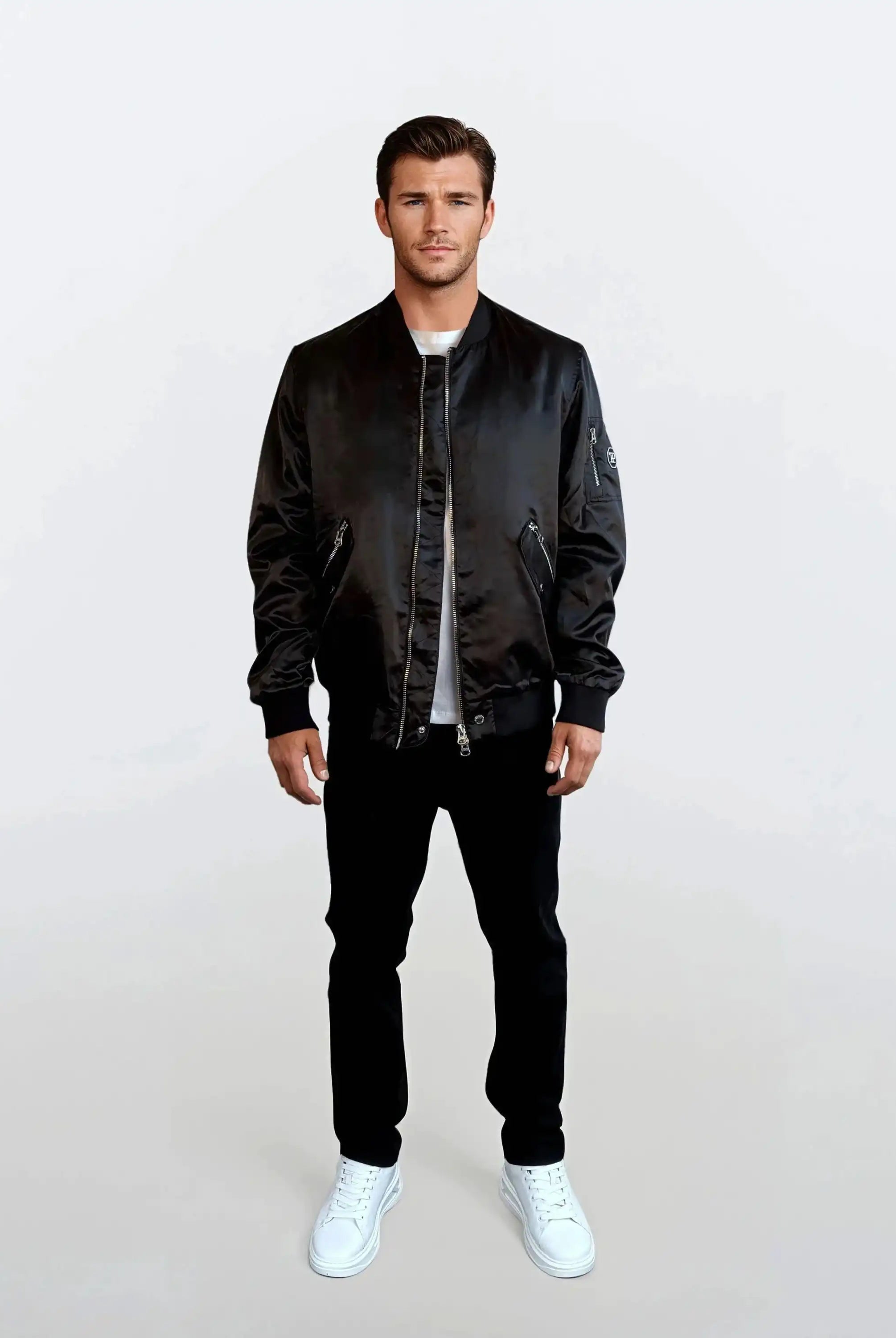 Ein Mann blickt vor einem weißen Hintergrund nach vorne und trägt einen urbanen Style mit einer trueprodigy Klassische Herren Bomber Jacke mit glänzender Oberfläche, einem weißen T-Shirt, einer schwarzen Hose und weißen Turnschuhen. Seine Arme sind entspannt an seinen Seiten.