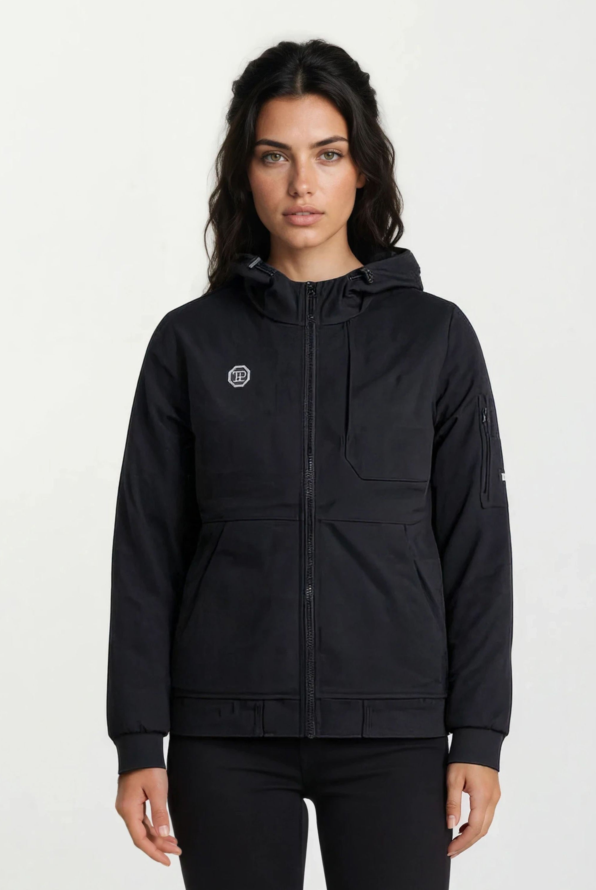 Eine Person mit langen dunklen Haaren trägt eine Trueprodigy Leichte Herrenjacke mit Brust- und Ärmelbadge, die mit einem Frontreißverschluss und mehreren Taschen versehen ist, vor einem schlichten hellen Hintergrund.