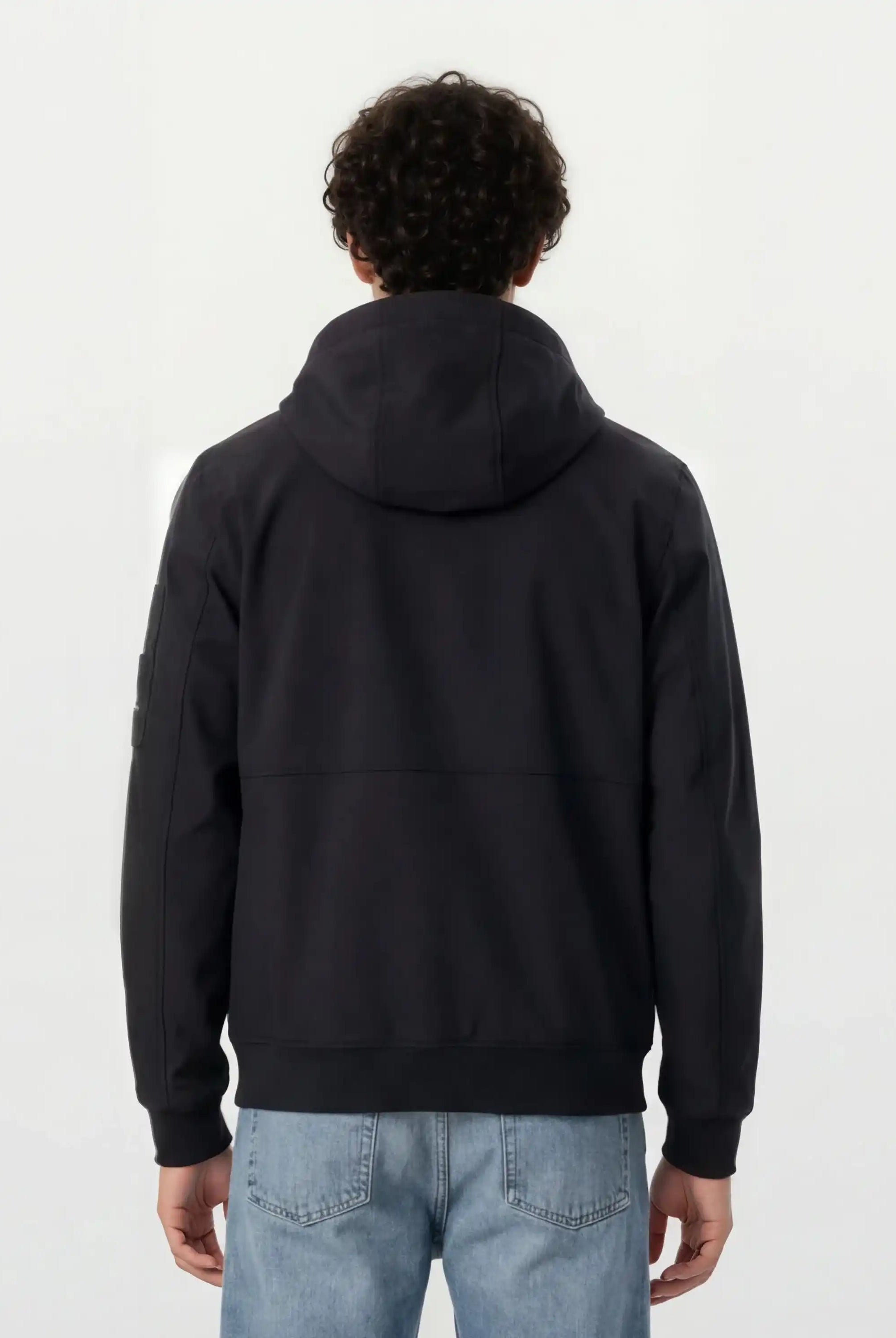 Eine Person mit lockigem Haar wird von hinten gezeigt, die eine Trueprodigy Leichte Herrenjacke mit Brust- und Ärmelbadge und hellblaue Jeans trägt und vor einem schlichten, hellen Hintergrund steht.