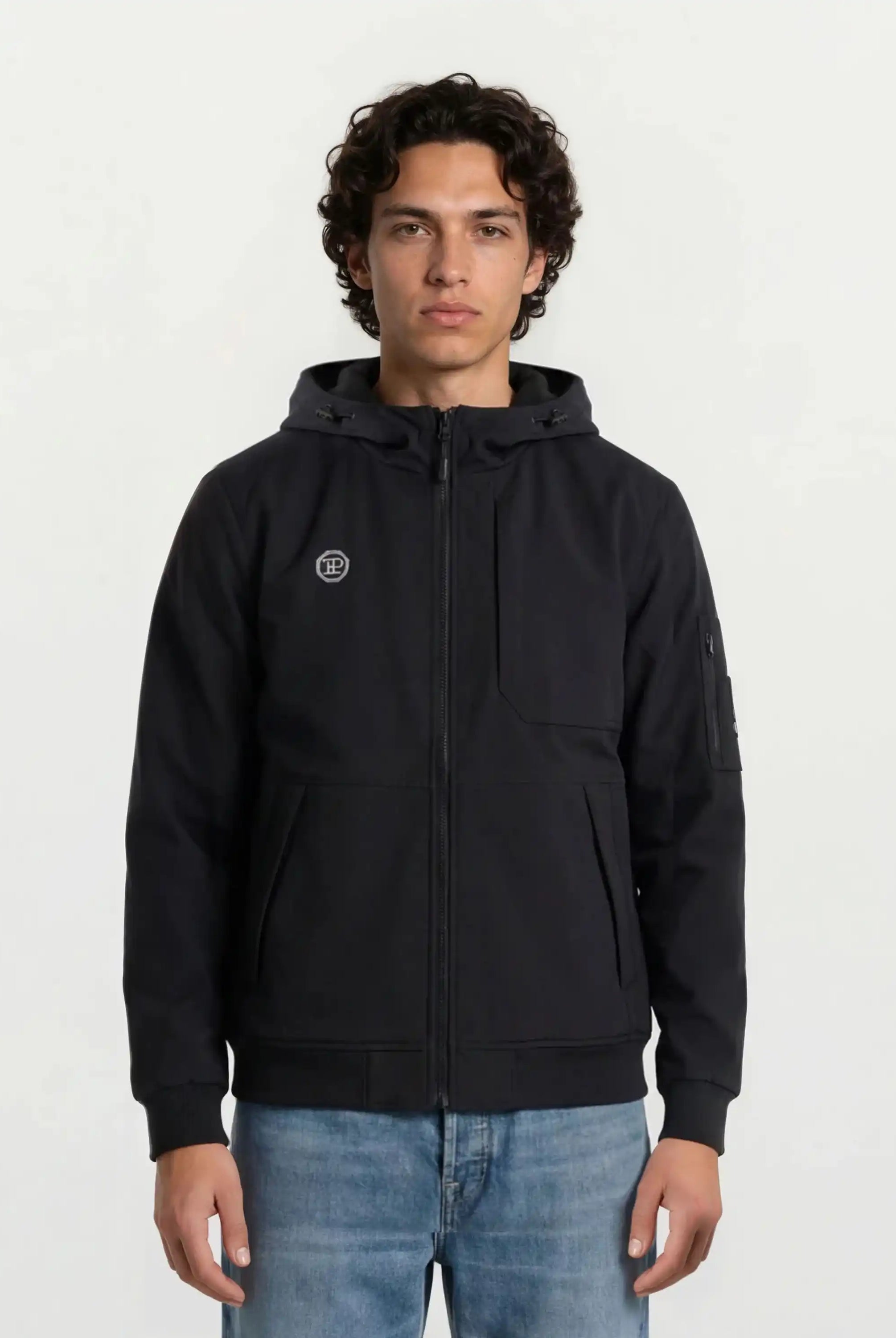 Eine Person mit lockigem Haar steht vor einem schlichten, hellen Hintergrund, trägt eine schwarze Leichte Herrenjacke mit Brust- und Ärmelbadge von trueprodigy und blaue Jeans und blickt mit neutralem Blick in die Kamera.