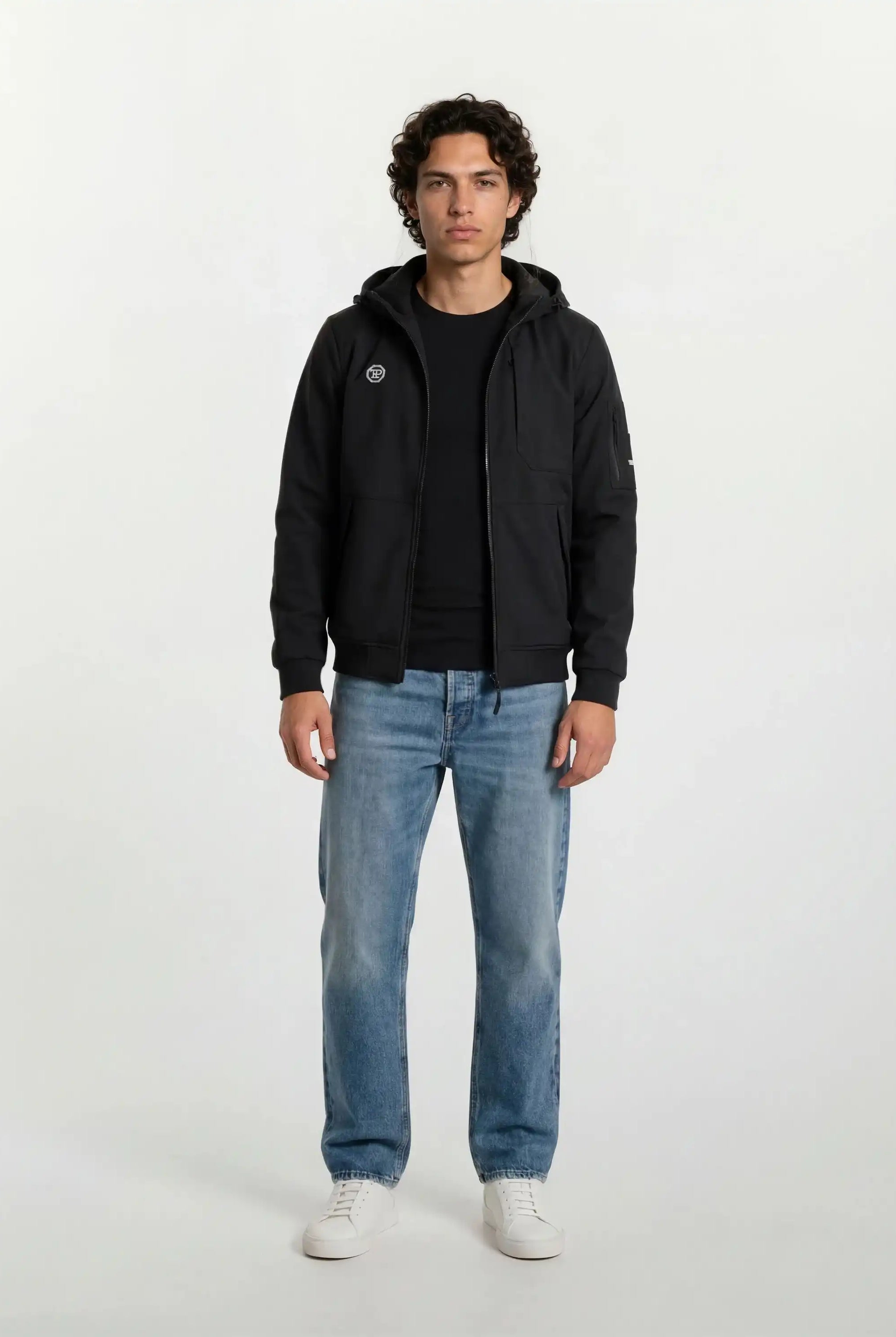 Eine Person steht mit Blick nach vorne in einer Trueprodigy Leichte Herrenjacke mit Brust- und Ärmelbadge, schwarzem T-Shirt, blauen Jeans und weißen Turnschuhen vor einem schlichten, hellen Hintergrund.