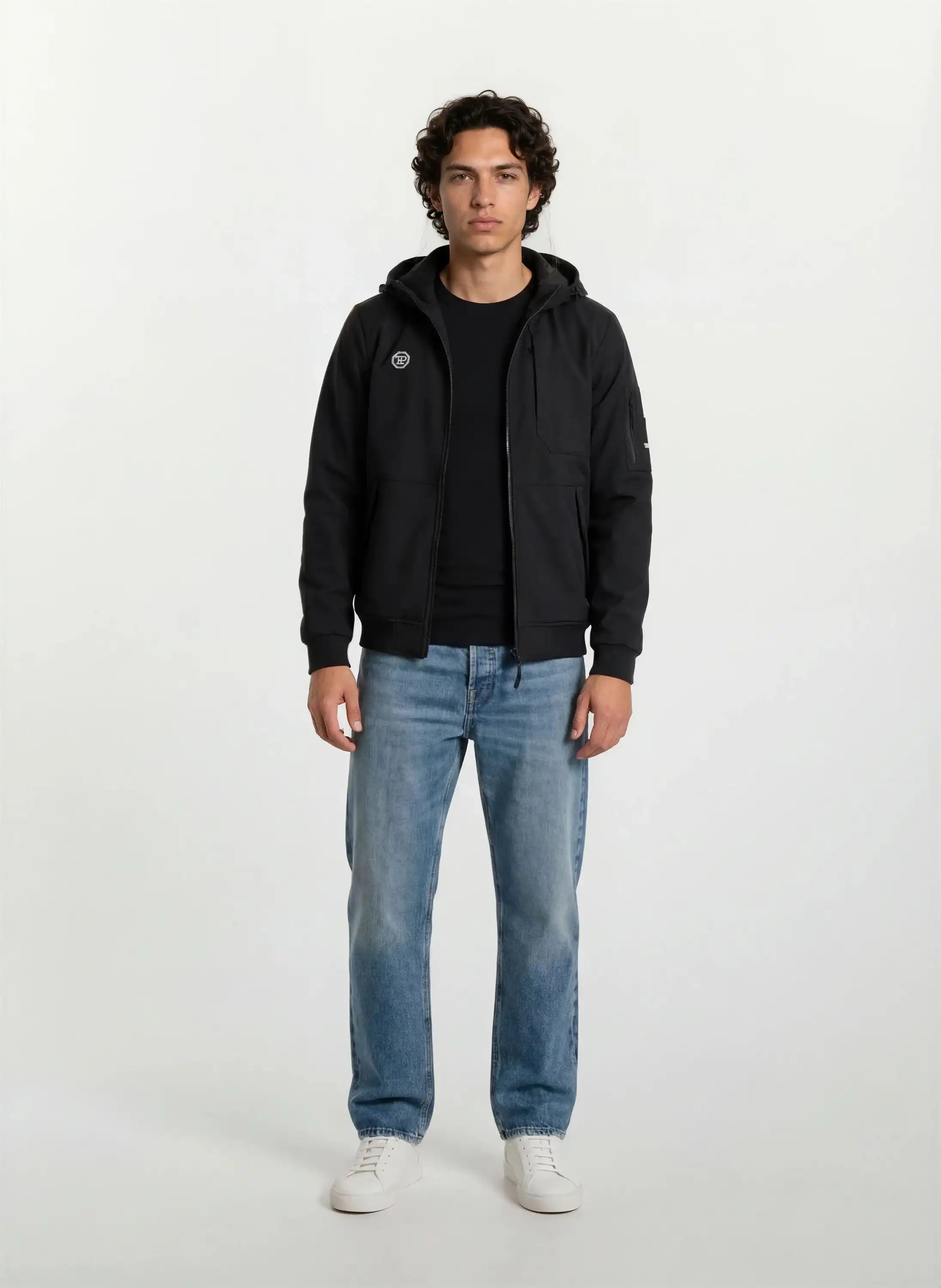 Eine Person steht mit Blick nach vorne in einer Trueprodigy Leichte Herrenjacke mit Brust- und Ärmelbadge, schwarzem T-Shirt, blauen Jeans und weißen Turnschuhen vor einem schlichten, hellen Hintergrund.