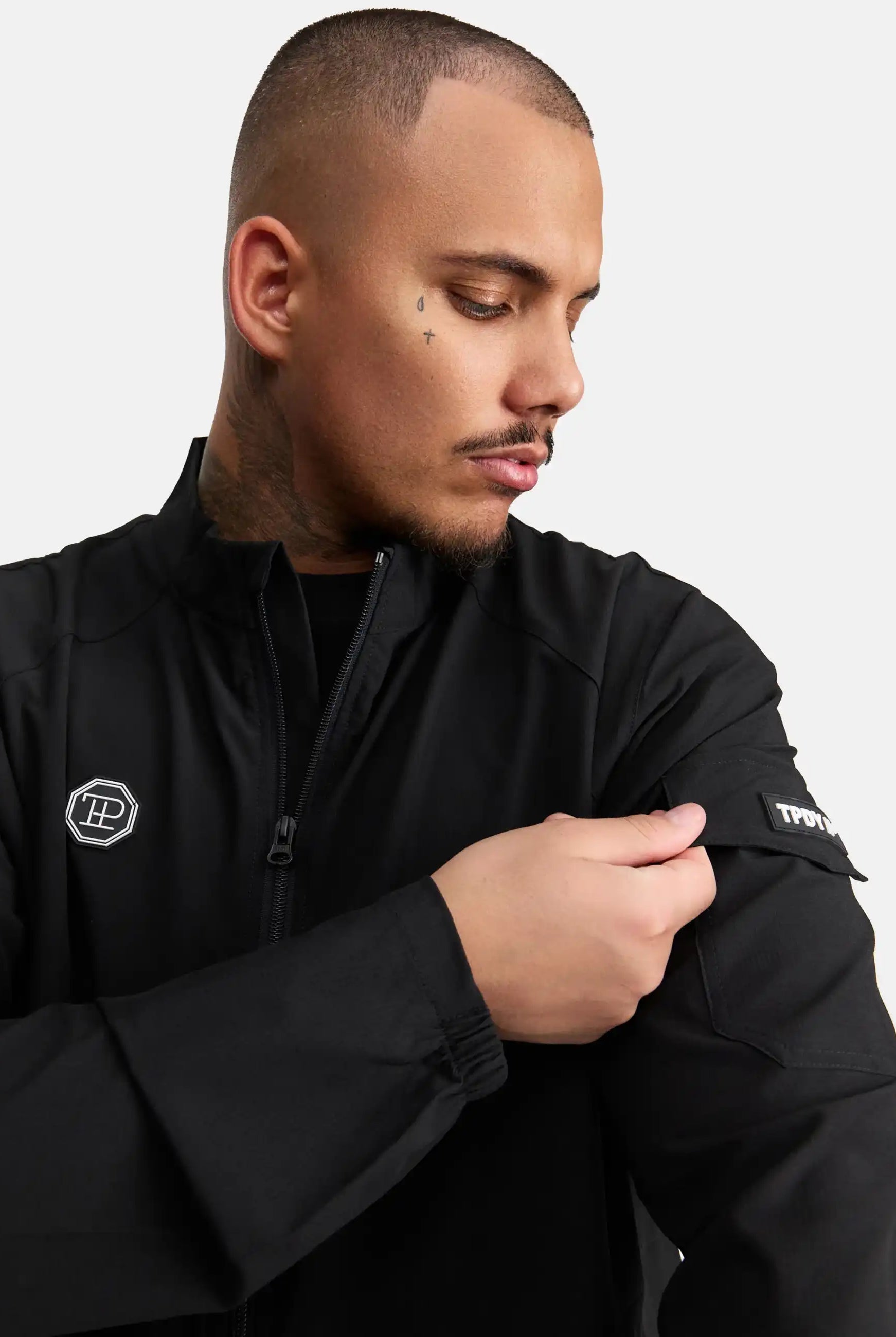 Ein Mann mit rasiertem Haarschnitt und Nackentattoos trägt die trueprodigy Henry Leichte Sportjacke mit Stehkragen in Schwarz und blickt auf seinen linken Arm, während er einen Aufnäher am Ärmel zurechtrückt. Der Hintergrund ist einfarbig weiß.