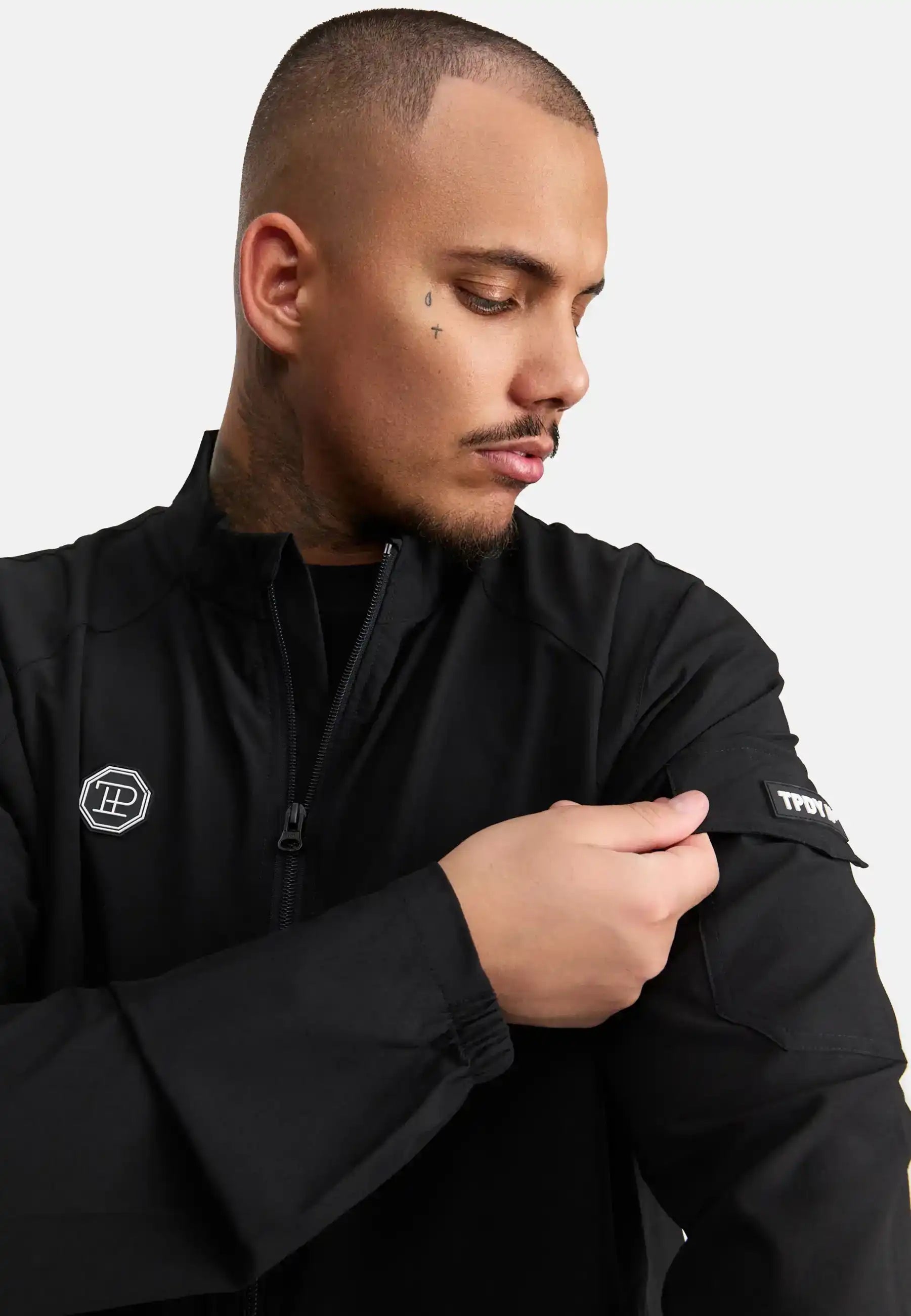 Ein Mann mit rasiertem Haarschnitt und Nackentattoos trägt die trueprodigy Henry Leichte Sportjacke mit Stehkragen in Schwarz und blickt auf seinen linken Arm, während er einen Aufnäher am Ärmel zurechtrückt. Der Hintergrund ist einfarbig weiß.