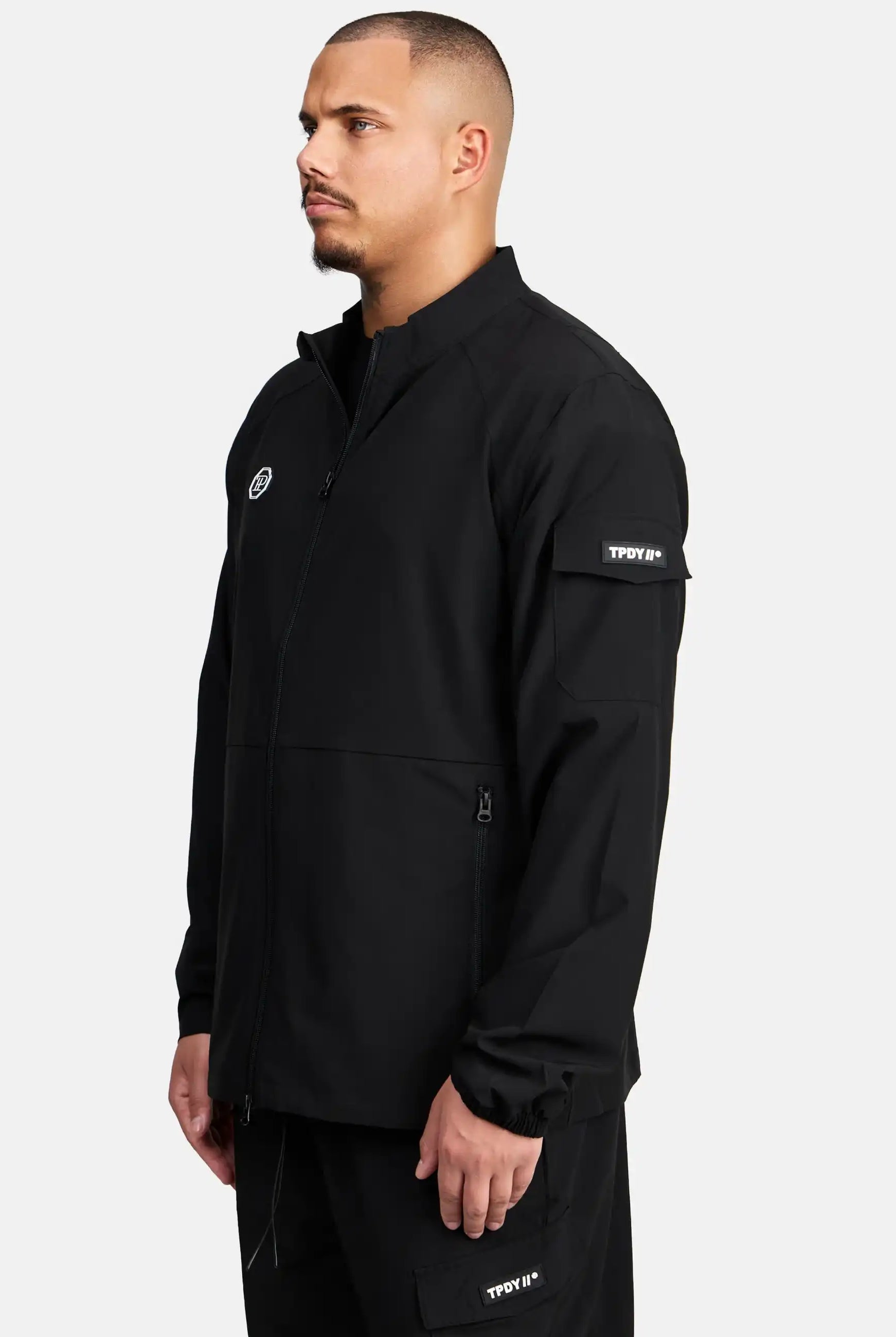 Ein Mann mit kurzen Haaren und gepflegtem Bart trägt die trueprodigy Henry leichte Sportjacke mit Stehkragen, Reißverschlusstaschen und Logo-Patches auf Brust sowie linkem Ärmel. Er steht vor hellem Hintergrund und blickt nach links.