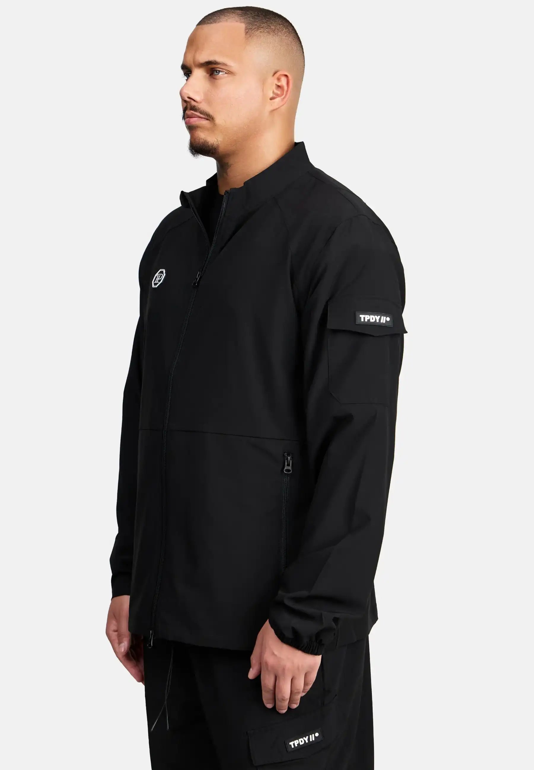 Ein Mann mit kurzen Haaren und gepflegtem Bart trägt die trueprodigy Henry leichte Sportjacke mit Stehkragen, Reißverschlusstaschen und Logo-Patches auf Brust sowie linkem Ärmel. Er steht vor hellem Hintergrund und blickt nach links.