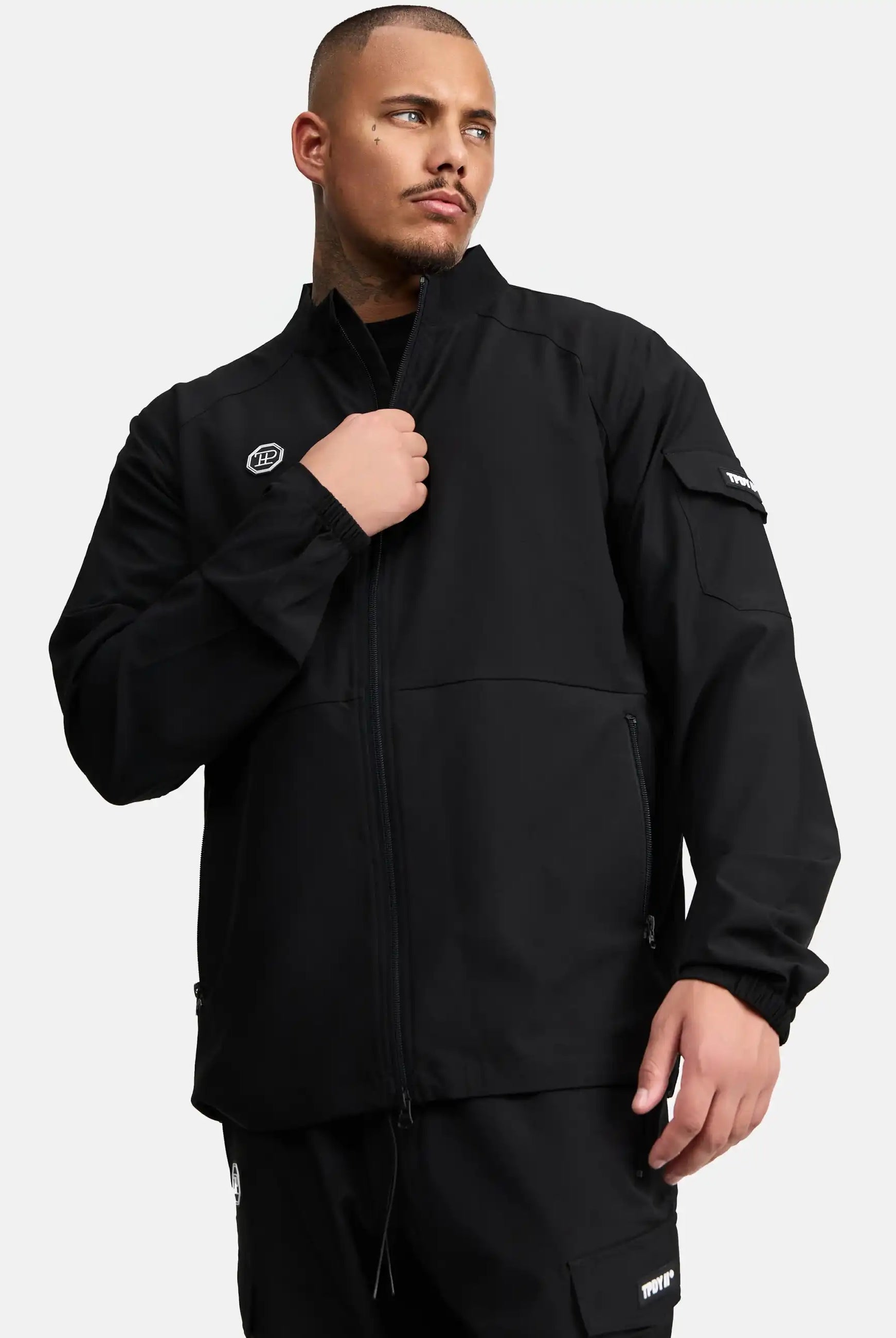 Ein Mann mit kurzem Haar und Bart trägt die Henry Leichte Sportjacke mit Stehkragen von trueprodigy und eine passende Hose. Er hält den Zipper der Jacke, während er seitlich blickt. Der Hintergrund ist schlicht weiß.