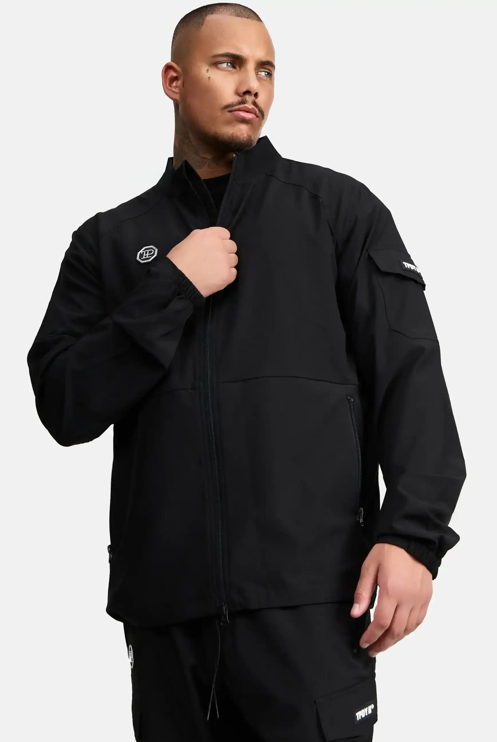 Ein Mann mit kurzem Haar und Bart trägt die Henry Leichte Sportjacke mit Stehkragen von trueprodigy und eine passende Hose. Er hält den Zipper der Jacke, während er seitlich blickt. Der Hintergrund ist schlicht weiß.