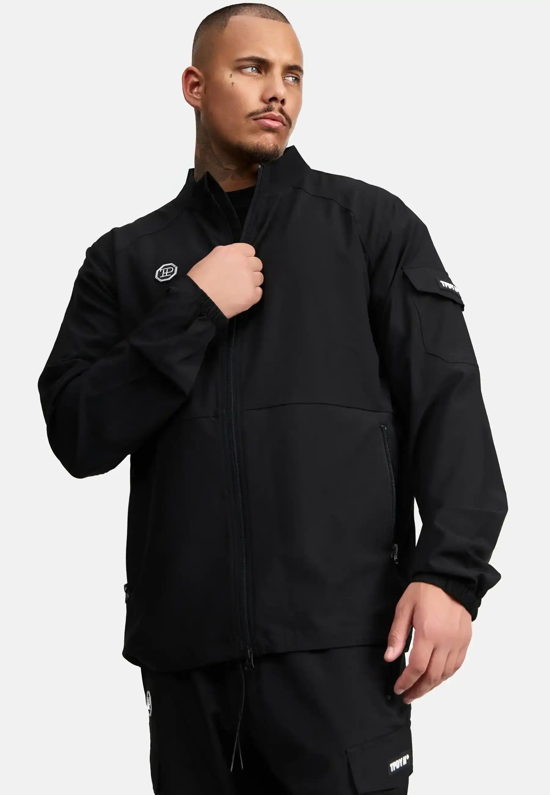 Ein Mann mit kurzem Haar und Bart trägt die Henry Leichte Sportjacke mit Stehkragen von trueprodigy und eine passende Hose. Er hält den Zipper der Jacke, während er seitlich blickt. Der Hintergrund ist schlicht weiß.