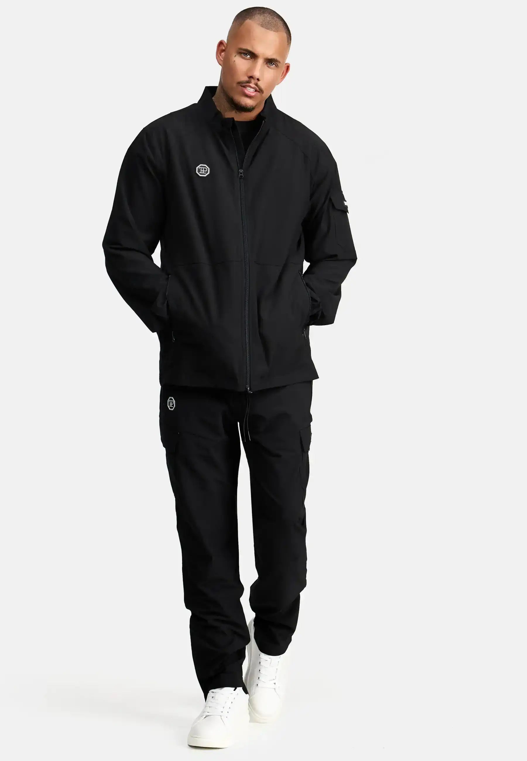 Ein Mann steht vor einem schlichten weißen Hintergrund und trägt die schwarze trueprodigy Henry Leichte Sportjacke mit Stehkragen und die dazu passende schwarze Cargohose mit weißen Logos sowie weiße Turnschuhe. Seine Hände stecken in den Hosentaschen, während er nach vorne blickt.