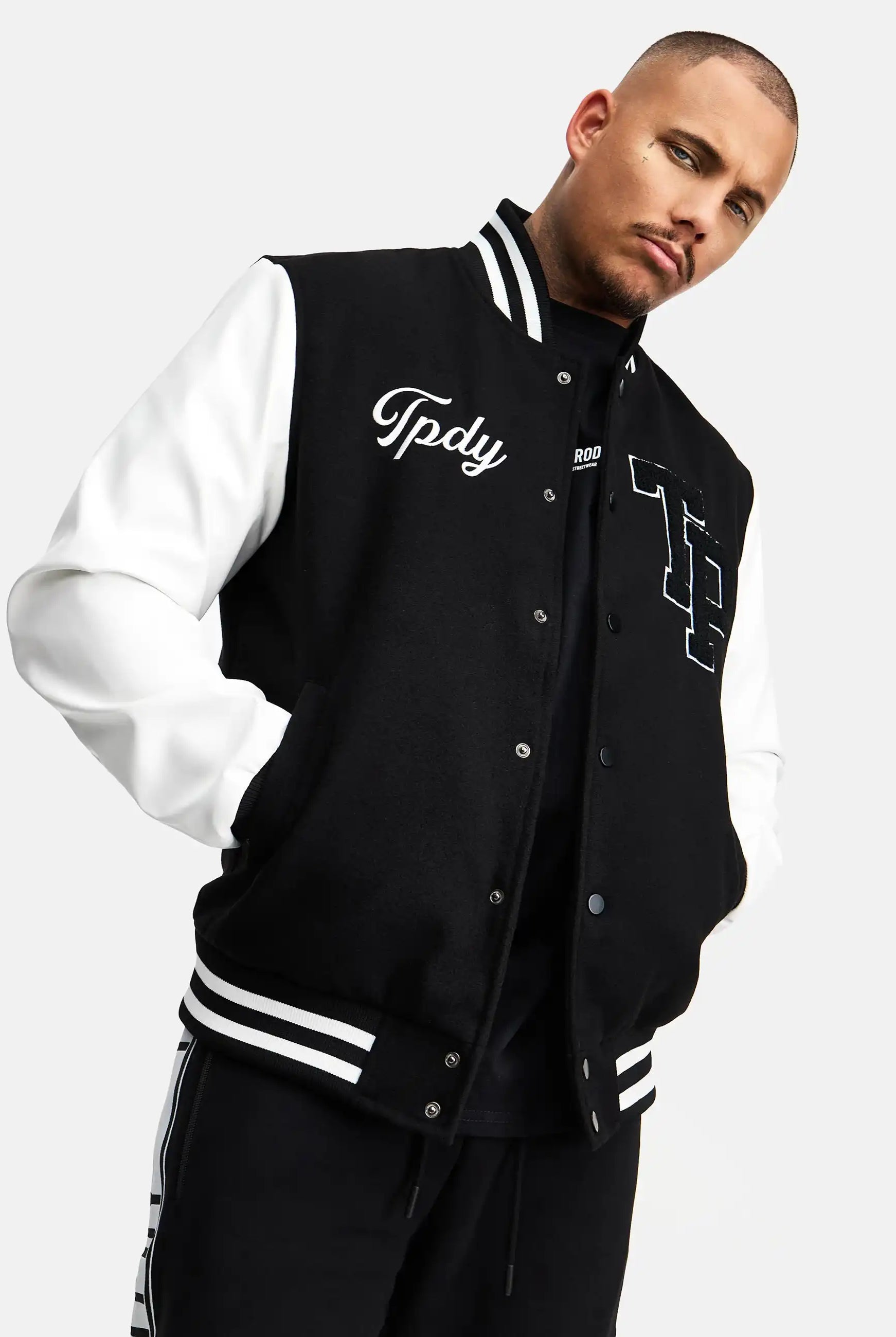 Ein Mann steht mit den Händen in den Taschen einer schwarz-weißen trueprodigy Chuck Retro Collage Jacke mit Logo, gestylt wie eine klassische College-Jacke mit gestickten Details. Er trägt eine schwarze Hose mit weißen Streifen und blickt in die Kamera.