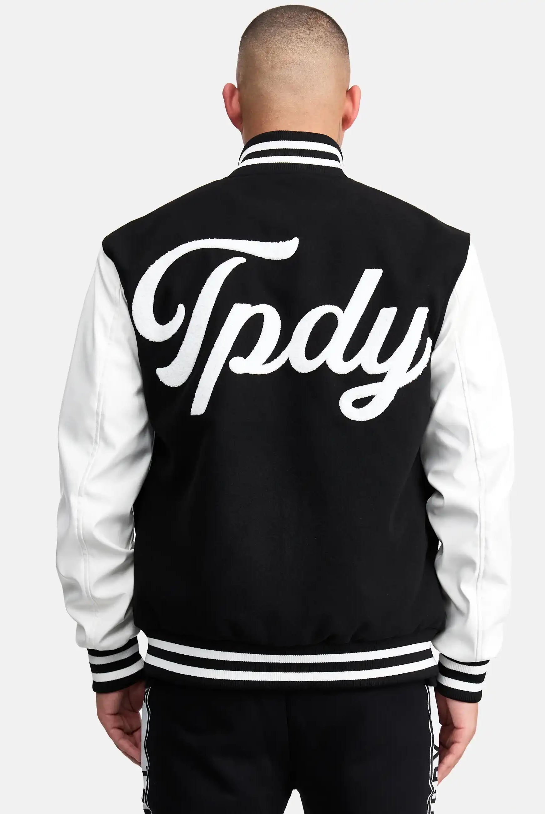Eine Person mit rasiertem Kopf ist von hinten zu sehen, die eine schwarz-weiße trueprodigy Chuck Retro Collage Jacke mit Logo trägt. Auf der Rückseite der Jacke steht in großen, kursiven weißen Buchstaben "Tpdy".