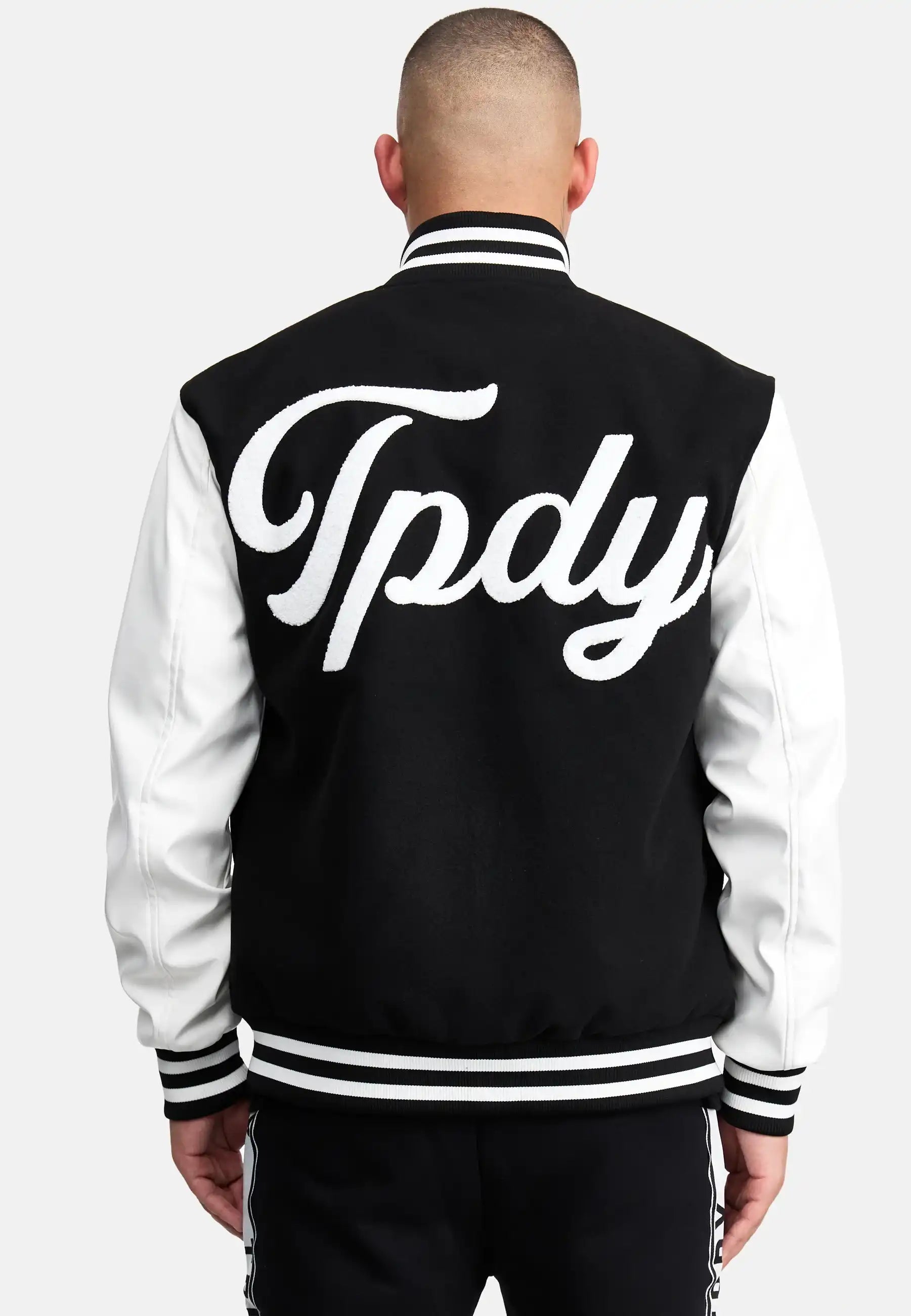 Eine Person mit rasiertem Kopf ist von hinten zu sehen, die eine schwarz-weiße trueprodigy Chuck Retro Collage Jacke mit Logo trägt. Auf der Rückseite der Jacke steht in großen, kursiven weißen Buchstaben "Tpdy".