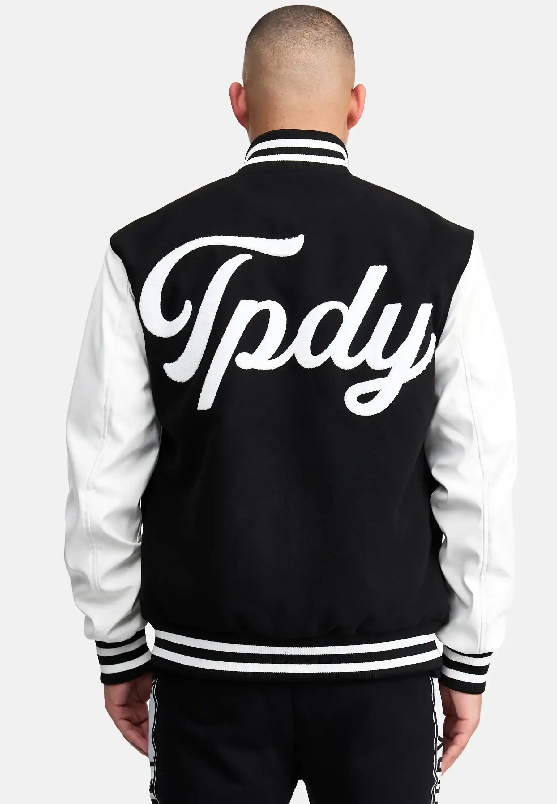 Eine Person mit rasiertem Kopf ist von hinten zu sehen, die eine schwarz-weiße trueprodigy Chuck Retro Collage Jacke mit Logo trägt. Auf der Rückseite der Jacke steht in großen, kursiven weißen Buchstaben "Tpdy".