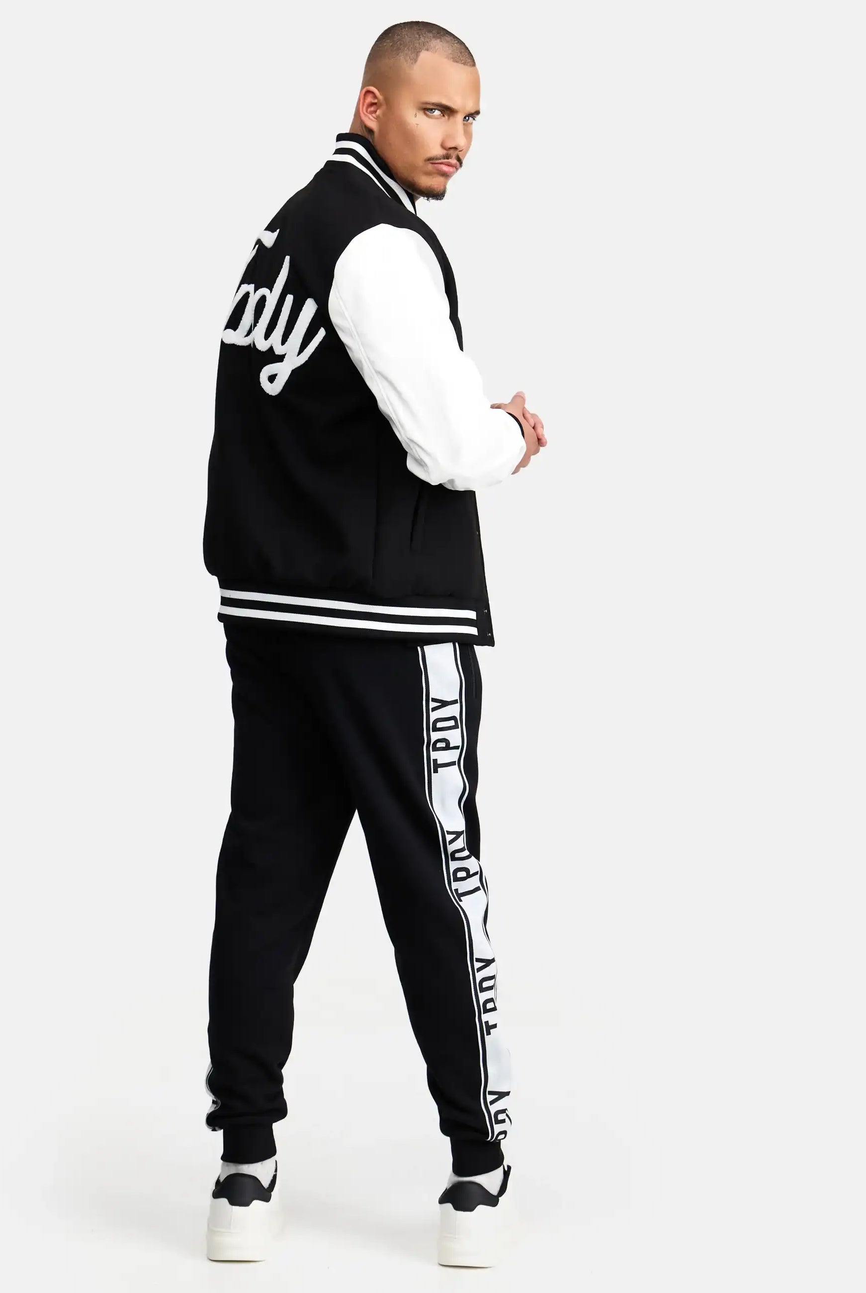 Ein Mann blickt nach hinten und wendet seinen Kopf der Kamera zu. Er trägt eine schwarz-weiße trueprodigy Chuck Retro Collage Jacke mit Logo, eine schwarze Trainingshose mit weißen Seitenstreifen und weiße Turnschuhe.