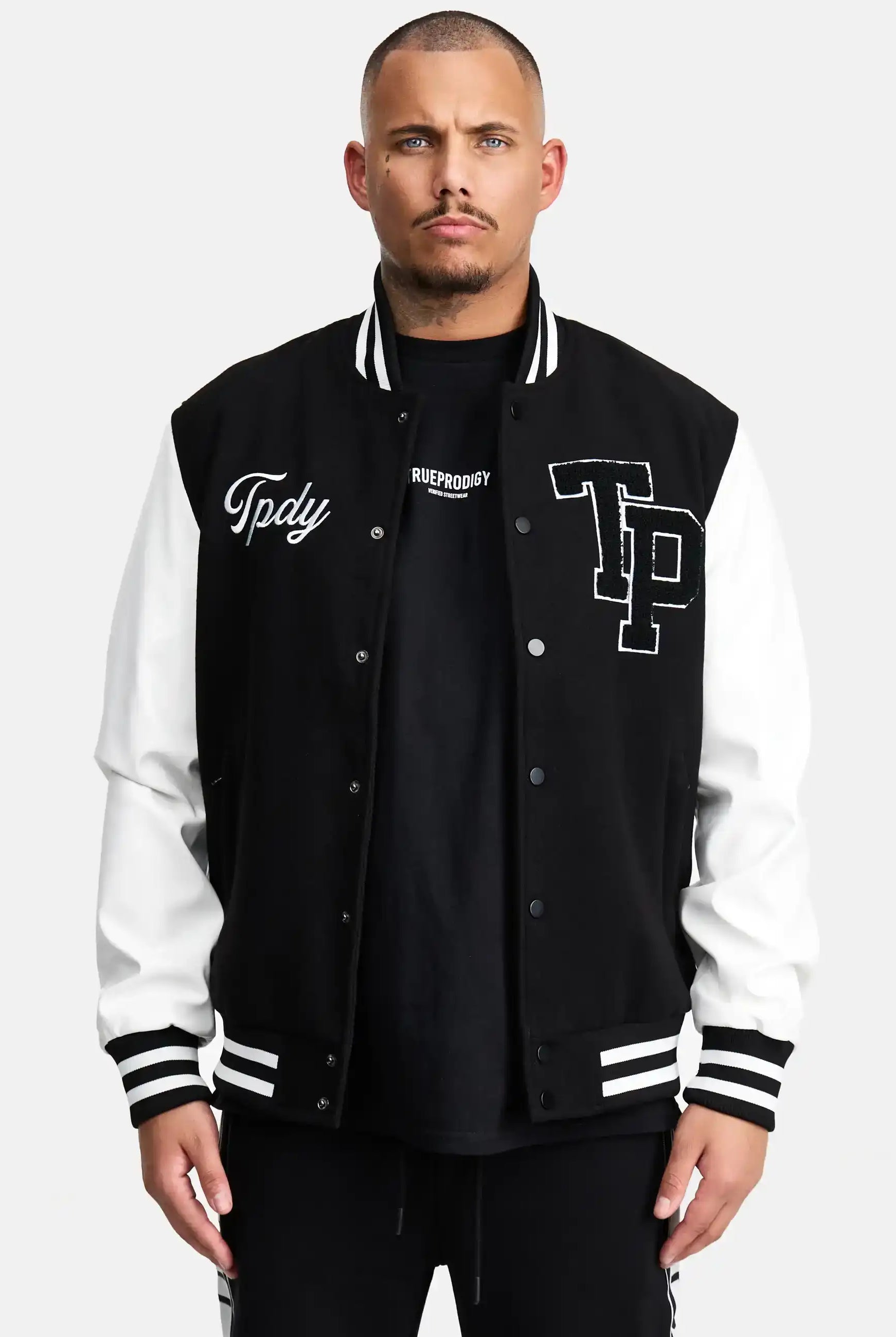 Ein Mann mit kurzen Haaren und neutralem Gesichtsausdruck steht in der trueprodigy Chuck Retro Collage Jacke mit Logo, schwarz-weiß, mit Tpdy- und TP-Logos über einem schwarzen Hemd und unterstreicht damit den unverkennbaren Stil der Marke.