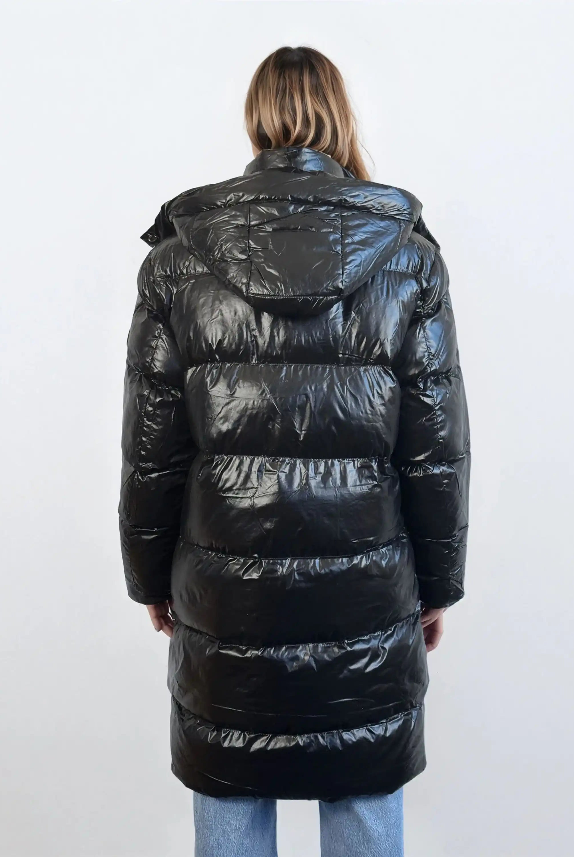 Eine Person mit schulterlangem Haar wird von hinten gezeigt, die eine glänzende trueprodigy Damen Winterjacke mit abnehmbarer Kapuze im Regular Fit trägt, gepaart mit hellblauen Jeans. Der Hintergrund ist einfarbig und hell gefärbt.