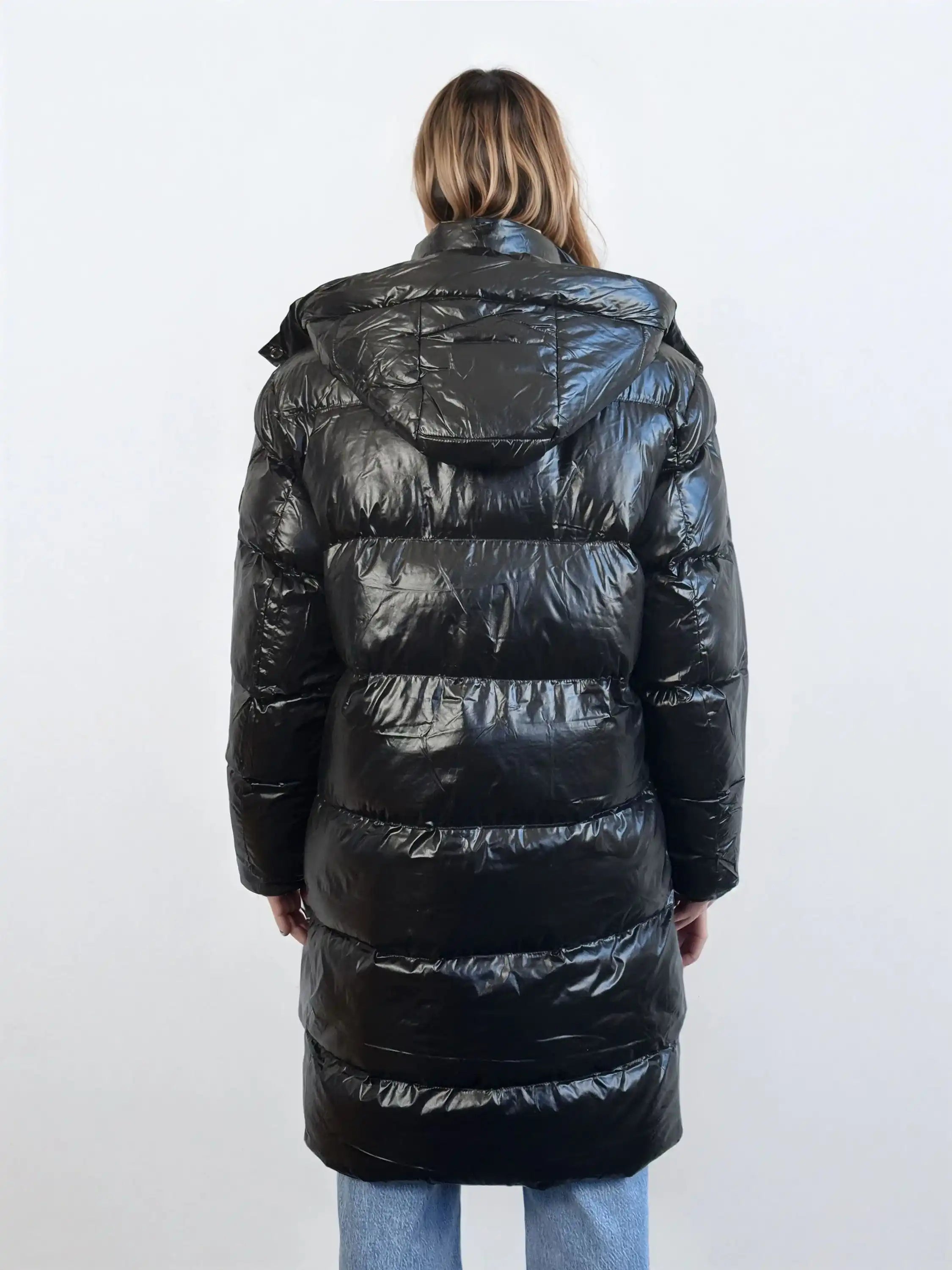 Eine Person mit schulterlangem Haar wird von hinten gezeigt, die eine glänzende trueprodigy Damen Winterjacke mit abnehmbarer Kapuze im Regular Fit trägt, gepaart mit hellblauen Jeans. Der Hintergrund ist einfarbig und hell gefärbt.