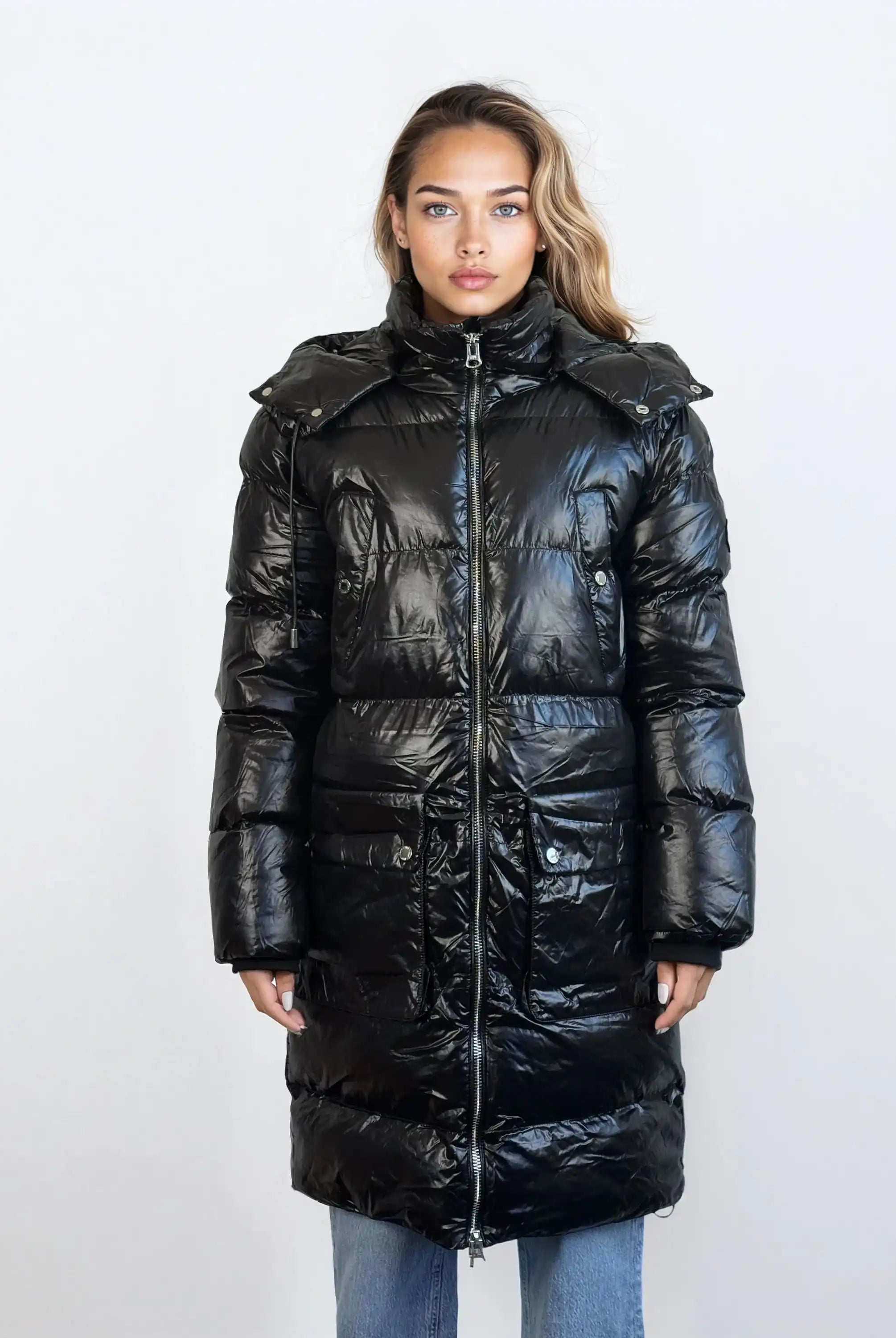 Eine Frau steht mit Blick nach vorne in einer glänzenden schwarzen trueprodigy Damen Winterjacke mit abnehmbarer Kapuze im Regular Fit mit Reißverschluss und hohem Kragen. Sie trägt eine blaue Jeans, die Hände an den Seiten, vor einem schlichten hellen Hintergrund.