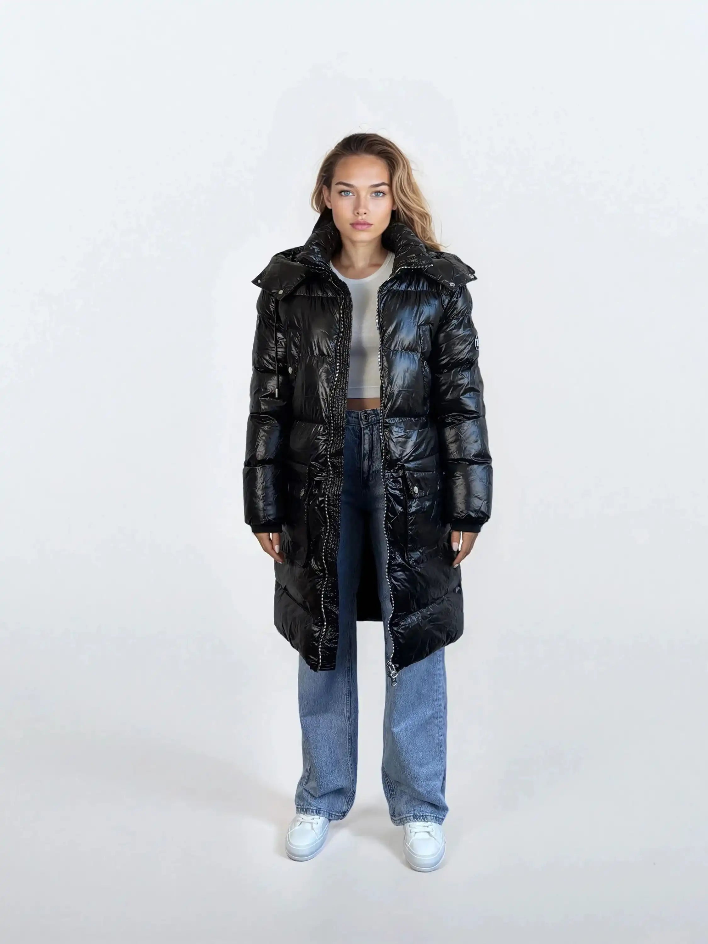 Eine Frau steht mit dem Gesicht nach vorne in einer glänzenden trueprodigy Damen Winterjacke mit abnehmbarer Kapuze im Regular Fit über einem weißen Crop Top, Blue Jeans und weißen Turnschuhen vor einem schlichten hellen Hintergrund.