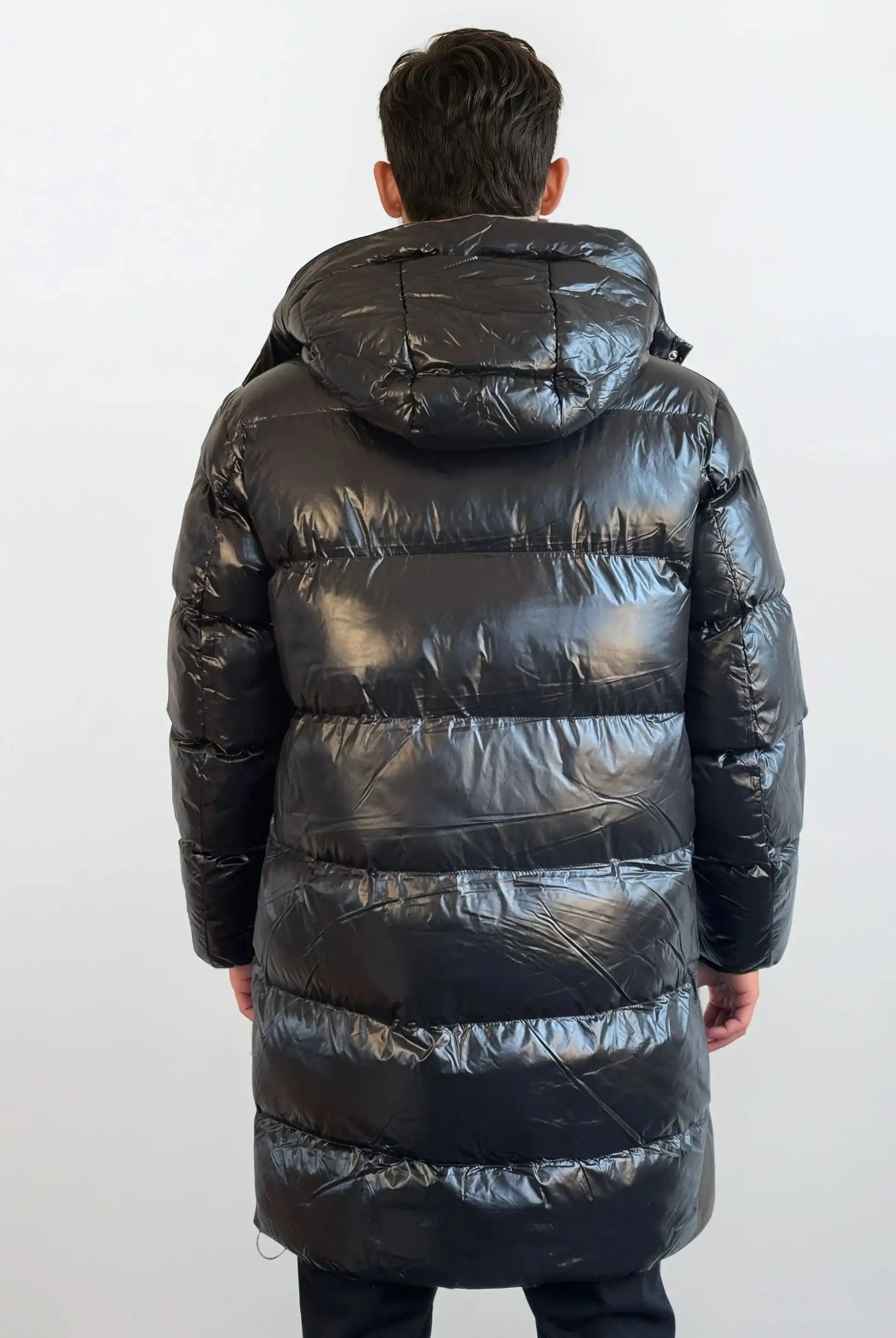 Eine Person mit kurzen dunklen Haaren steht mit dem Rücken zur Kamera und trägt eine glänzende trueprodigy Herren Winterjacke mit abnehmbarer Kapuze im Regular Fit in Schwarz, die in ihrer Länge bis über die Knie reicht, vor einem schlichten hellen Hintergrund.