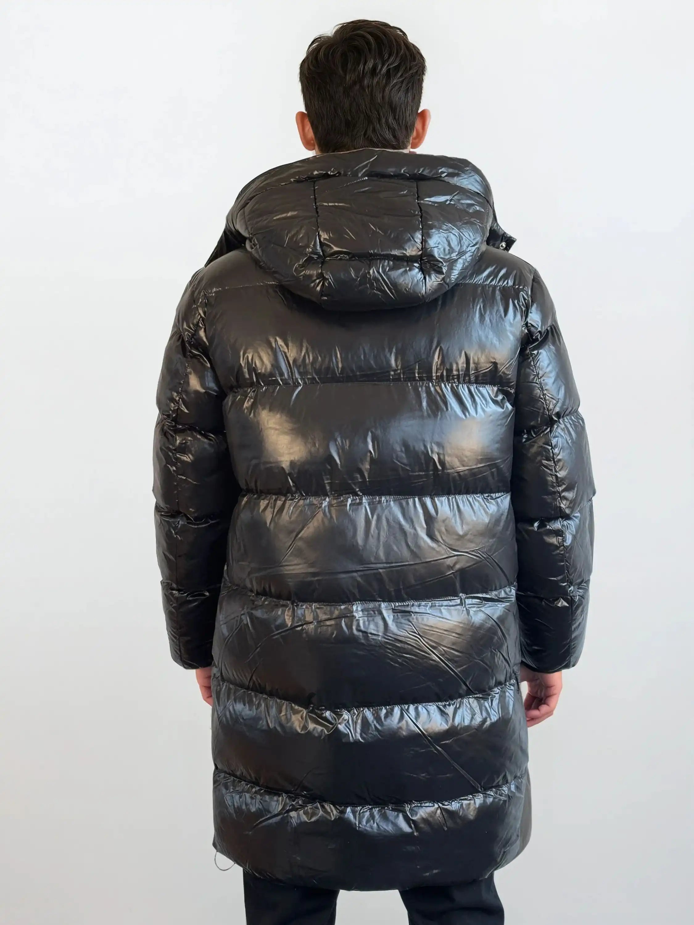 Eine Person mit kurzen dunklen Haaren steht mit dem Rücken zur Kamera und trägt eine glänzende trueprodigy Herren Winterjacke mit abnehmbarer Kapuze im Regular Fit in Schwarz, die in ihrer Länge bis über die Knie reicht, vor einem schlichten hellen Hintergrund.