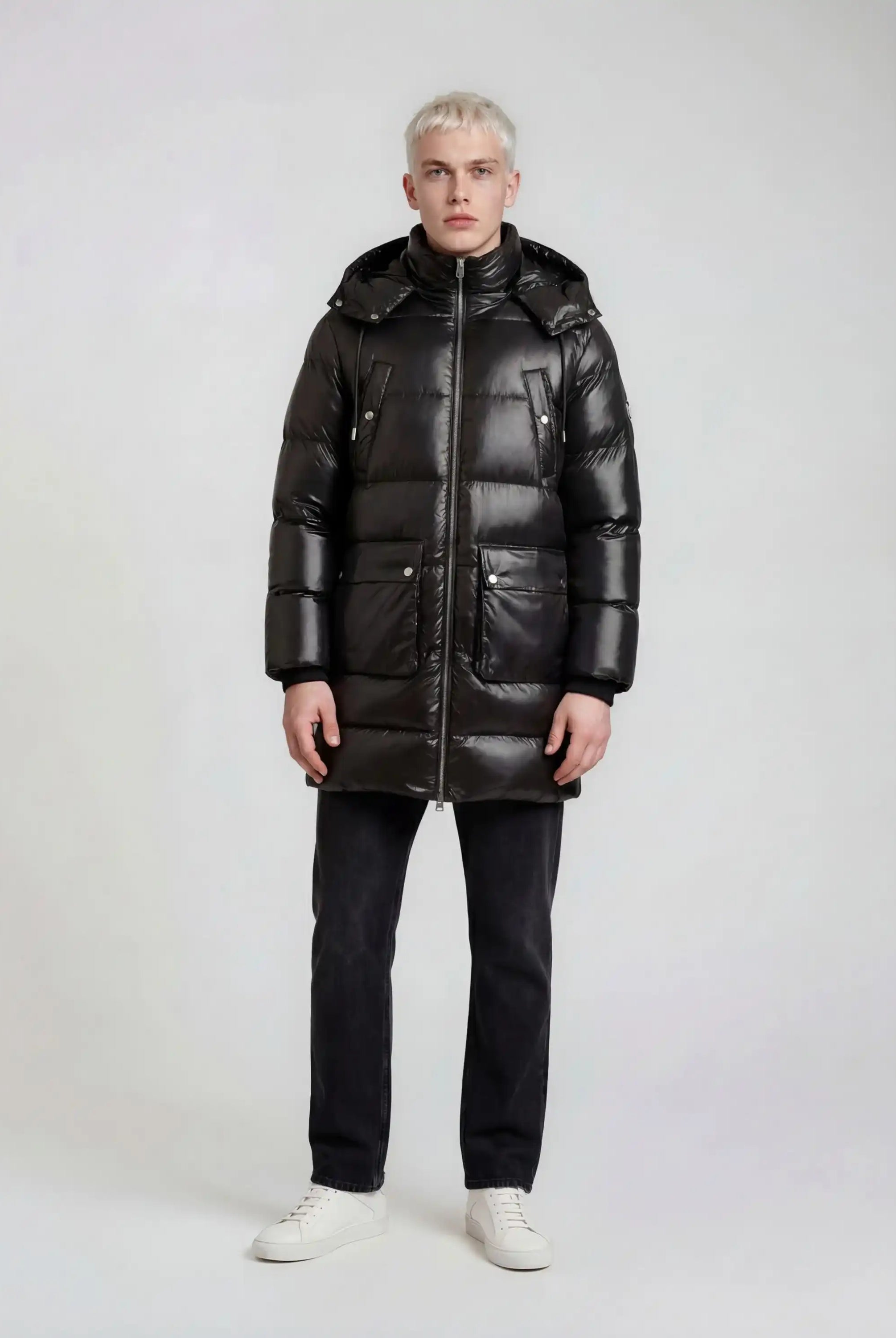 Eine Person mit kurzen blonden Haaren sieht nach vorne und trägt eine glänzende trueprodigy Herren Winterjacke mit abnehmbarer Kapuze im Regular Fit, eine schwarze Hose und weiße Turnschuhe vor einem schlichten hellen Hintergrund.