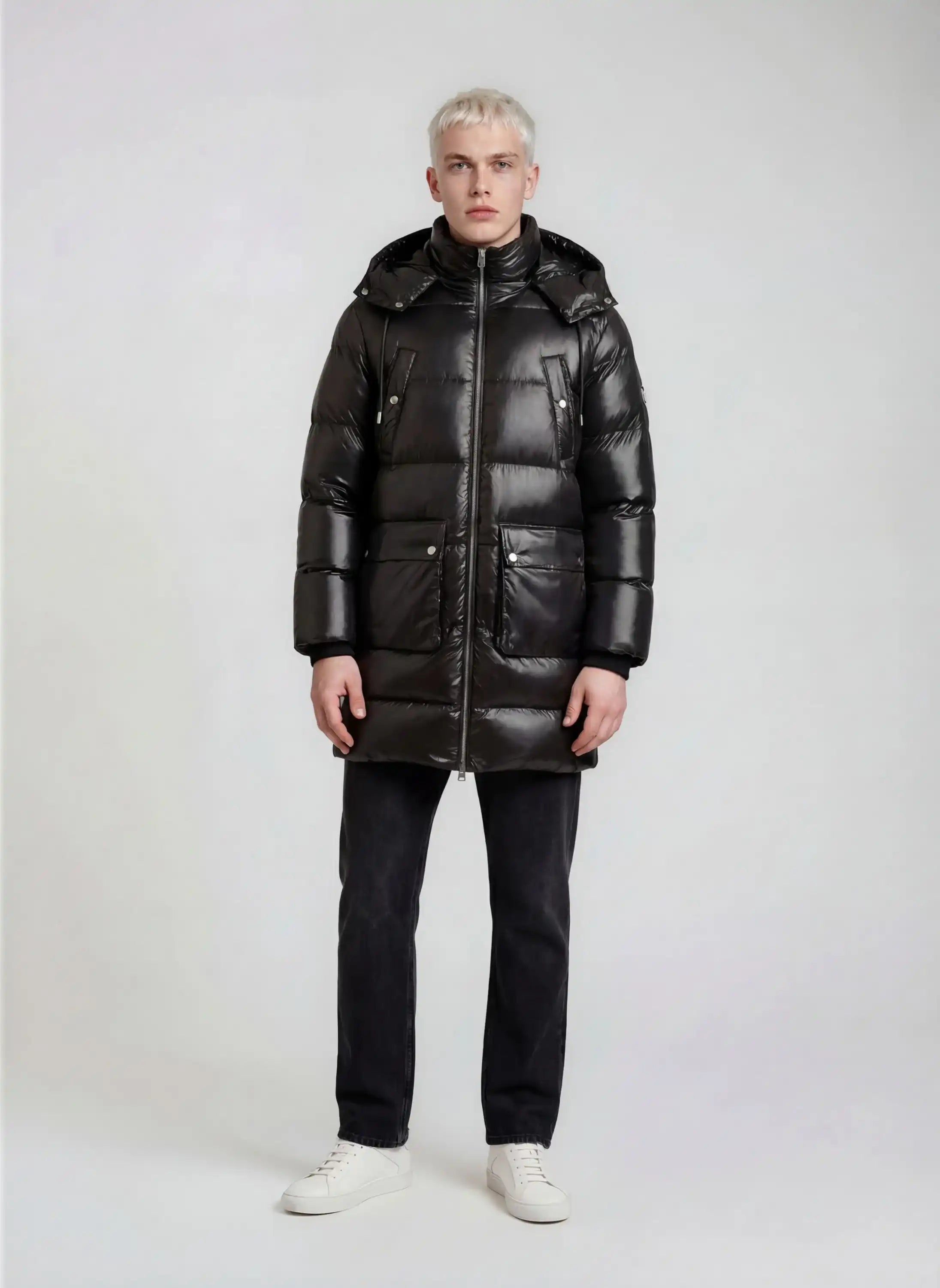 Eine Person mit kurzen blonden Haaren sieht nach vorne und trägt eine glänzende trueprodigy Herren Winterjacke mit abnehmbarer Kapuze im Regular Fit, eine schwarze Hose und weiße Turnschuhe vor einem schlichten hellen Hintergrund.