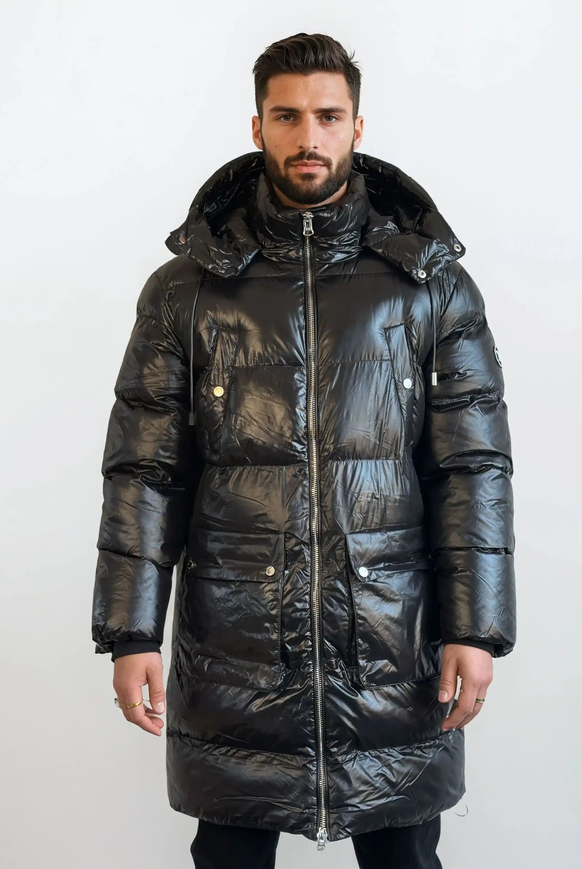 Ein Mann steht mit dem Gesicht nach vorne in einer glänzenden, langen schwarzen trueprodigy Herren Winterjacke mit abnehmbarer Kapuze im Regular Fit, die mit einem Frontreißverschluss und Druckknopftaschen ausgestattet ist, vor einem schlichten weißen Hintergrund.