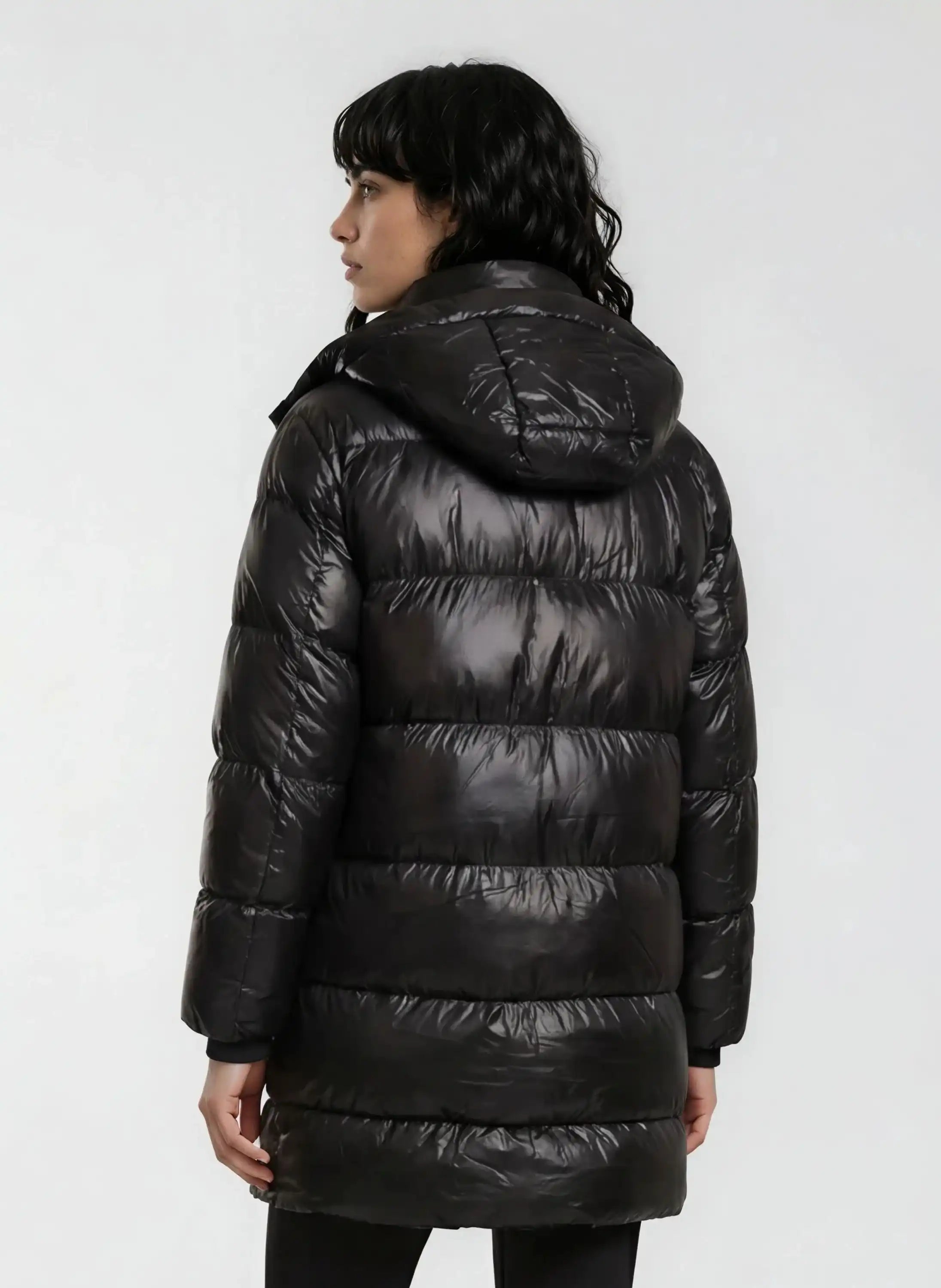 Eine Person mit dunklem, gewelltem Haar ist von hinten zu sehen, die eine glänzende trueprodigy Damen Winterjacke mit abnehmbarer Kapuze im Regular Fit trägt. Die glänzende Oberfläche der Jacke steht im Kontrast zu dem einfarbigen, hellen Hintergrund.