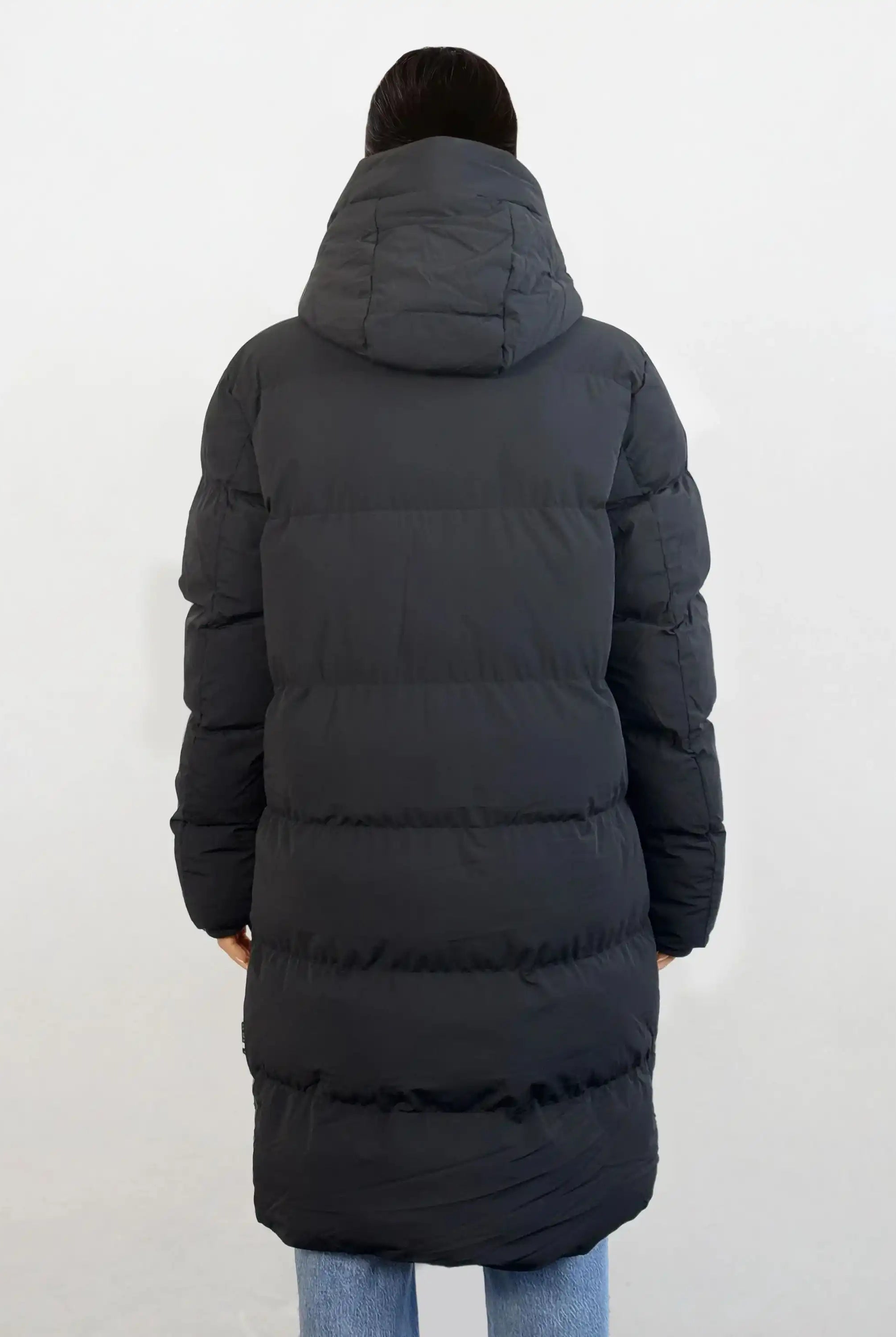 Man sieht eine Person von hinten, die den trueprodigy Damen Parka mit fester Kapuze im Regular Fit in Schwarz mit horizontaler Steppung trägt. Die wasserabweisende Jacke wird mit einer hellblauen Jeans vor einem schlichten, hellen Hintergrund kombiniert.