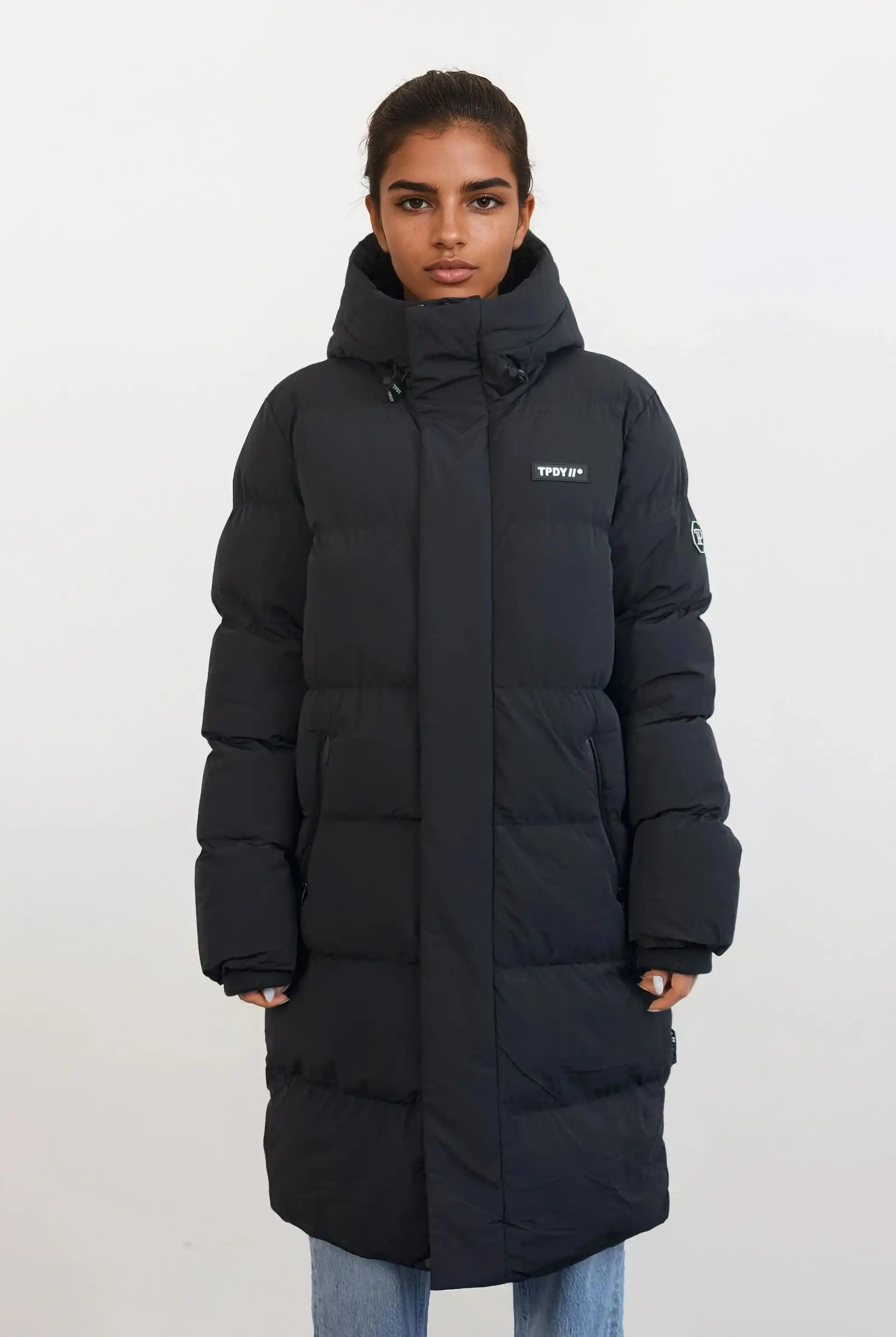 Eine Person steht mit dem Gesicht nach vorne und trägt den trueprodigy Damen Parka mit fester Kapuze im Regular Fit mit sichtbarem Brustlogo, gepaart mit einer hellblauen Jeans vor einem schlichten weißen Hintergrund.