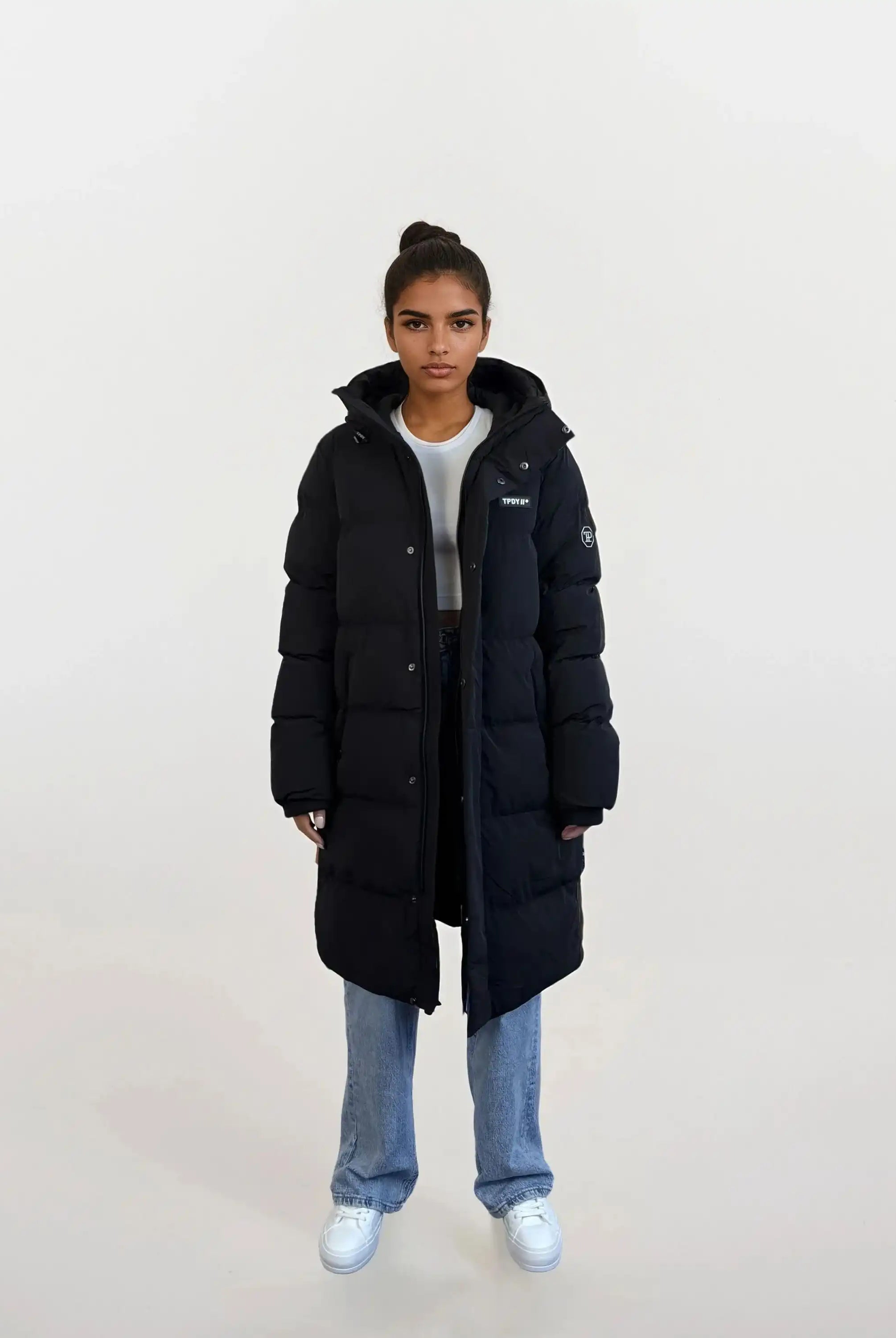 Eine Person trägt den trueprodigy Damen Parka mit fester Kapuze im Regular Fit über weißem Crop Top, blauer Jeans und weißen Sneakern. Die wasserabweisende Jacke wird vor hellem Hintergrund präsentiert, das Haar ist hochgebunden.