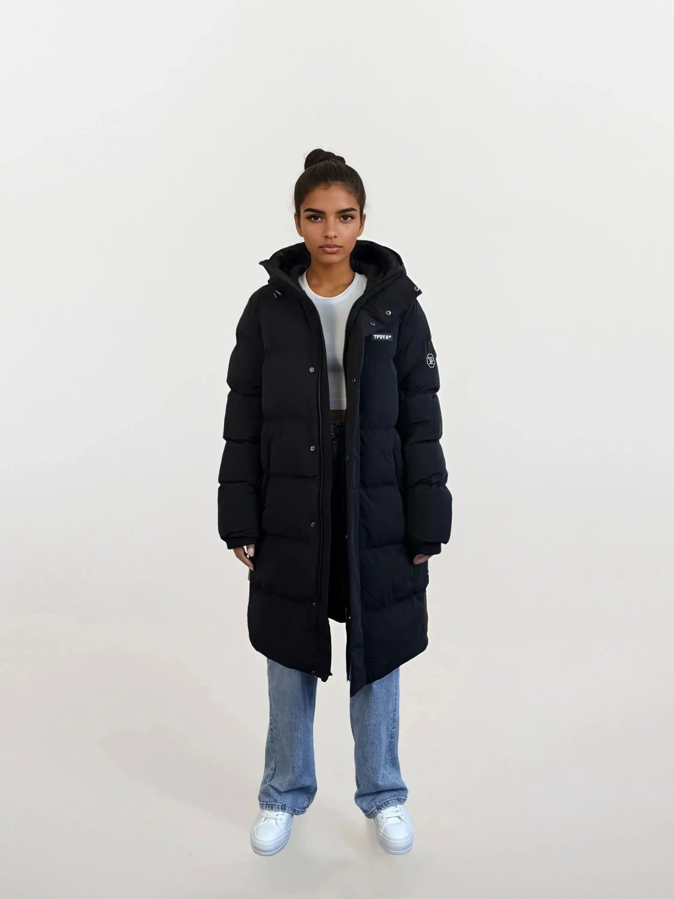 Eine Person trägt den trueprodigy Damen Parka mit fester Kapuze im Regular Fit über weißem Crop Top, blauer Jeans und weißen Sneakern. Die wasserabweisende Jacke wird vor hellem Hintergrund präsentiert, das Haar ist hochgebunden.