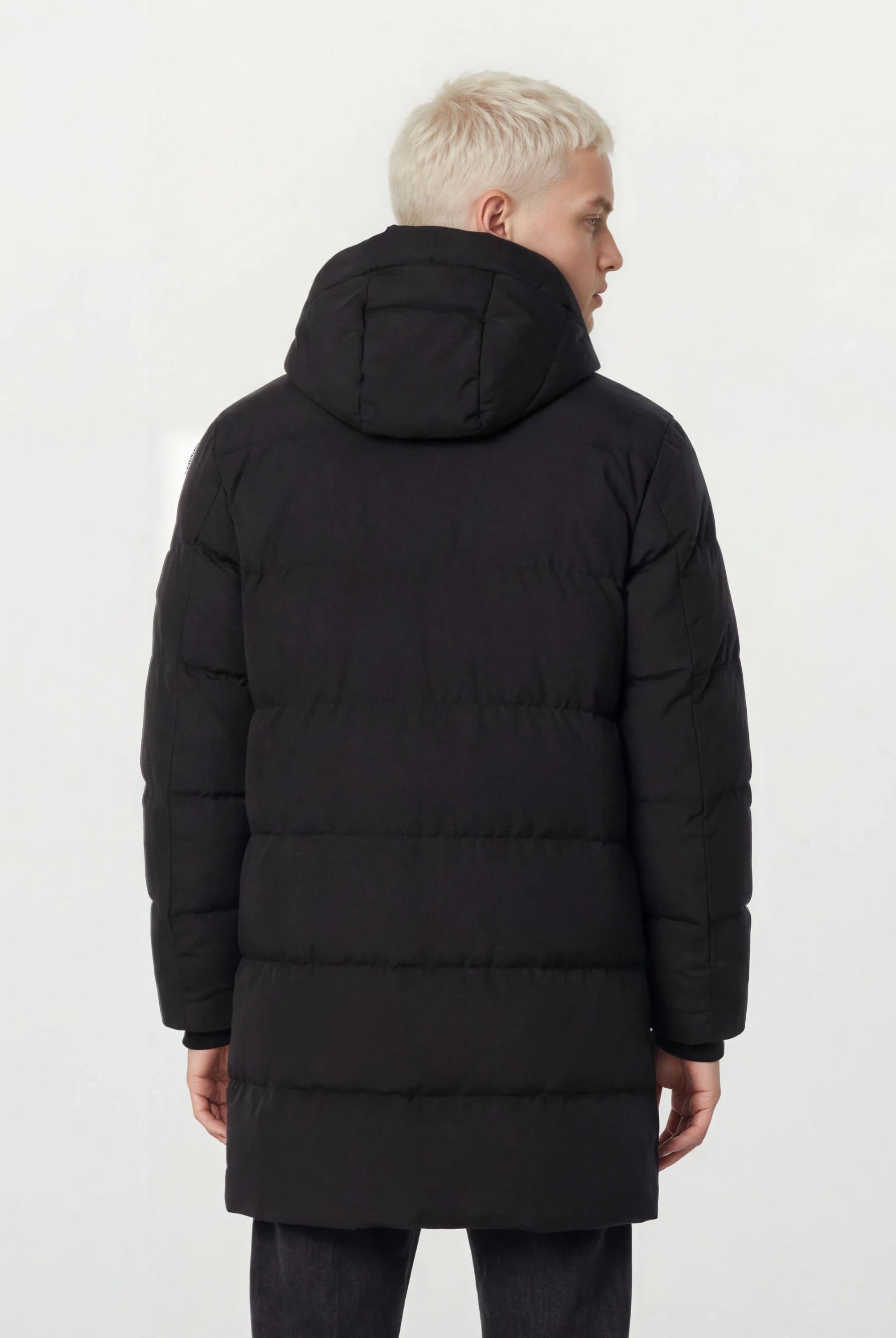 Man sieht eine Person mit kurzen, hellblonden Haaren von hinten, die den trueprodigy Herren Parka mit fester Kapuze im Regular Fit trägt. Der wasserabweisende schwarze Mantel hebt sich deutlich von dem schlichten, hellen Hintergrund ab.