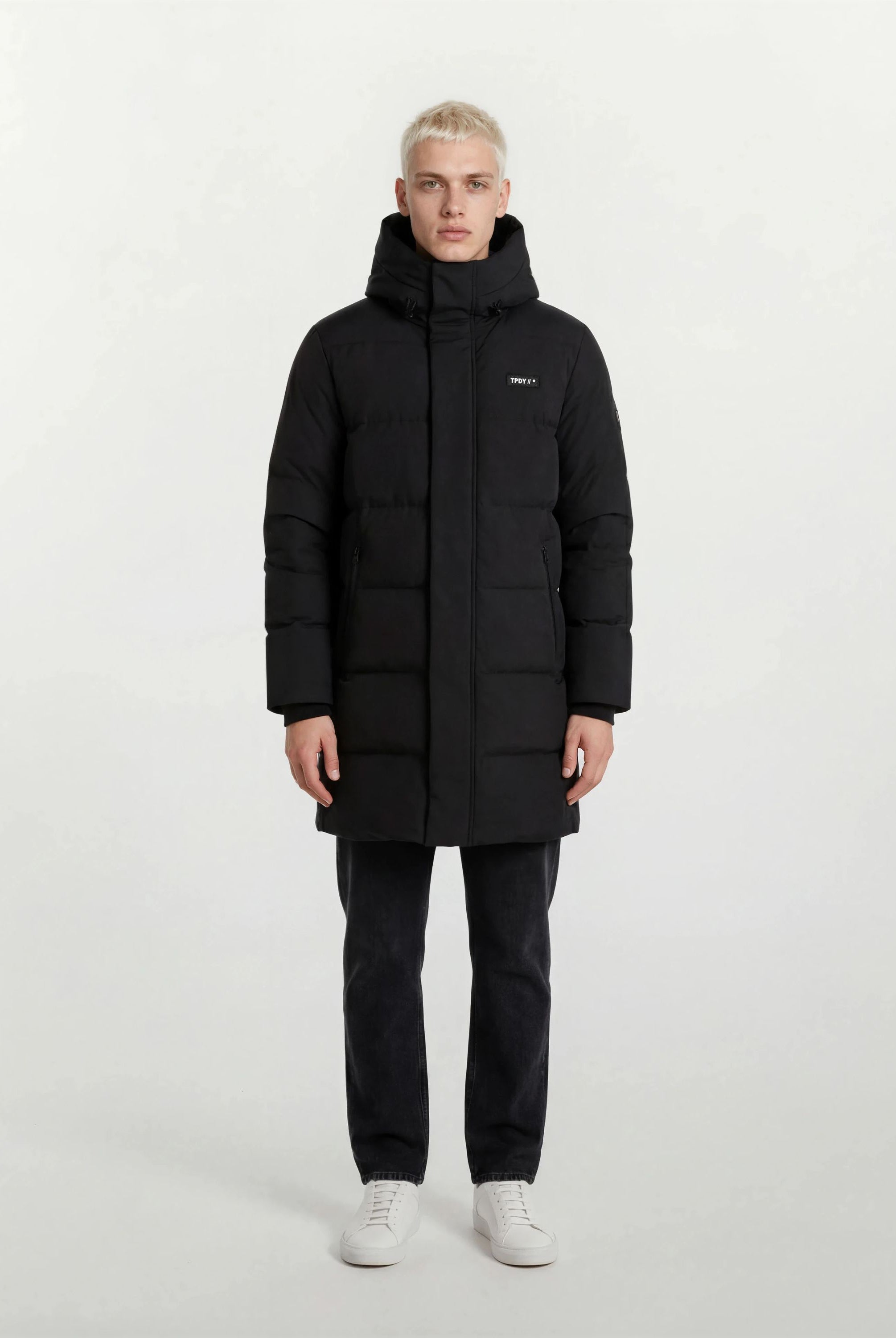 Eine Person mit kurzen blonden Haaren steht mit dem Gesicht nach vorne und trägt den trueprodigy Herren Parka mit fester Kapuze im Regular Fit, eine dunkle Hose und weiße Turnschuhe vor einem schlichten hellen Hintergrund.