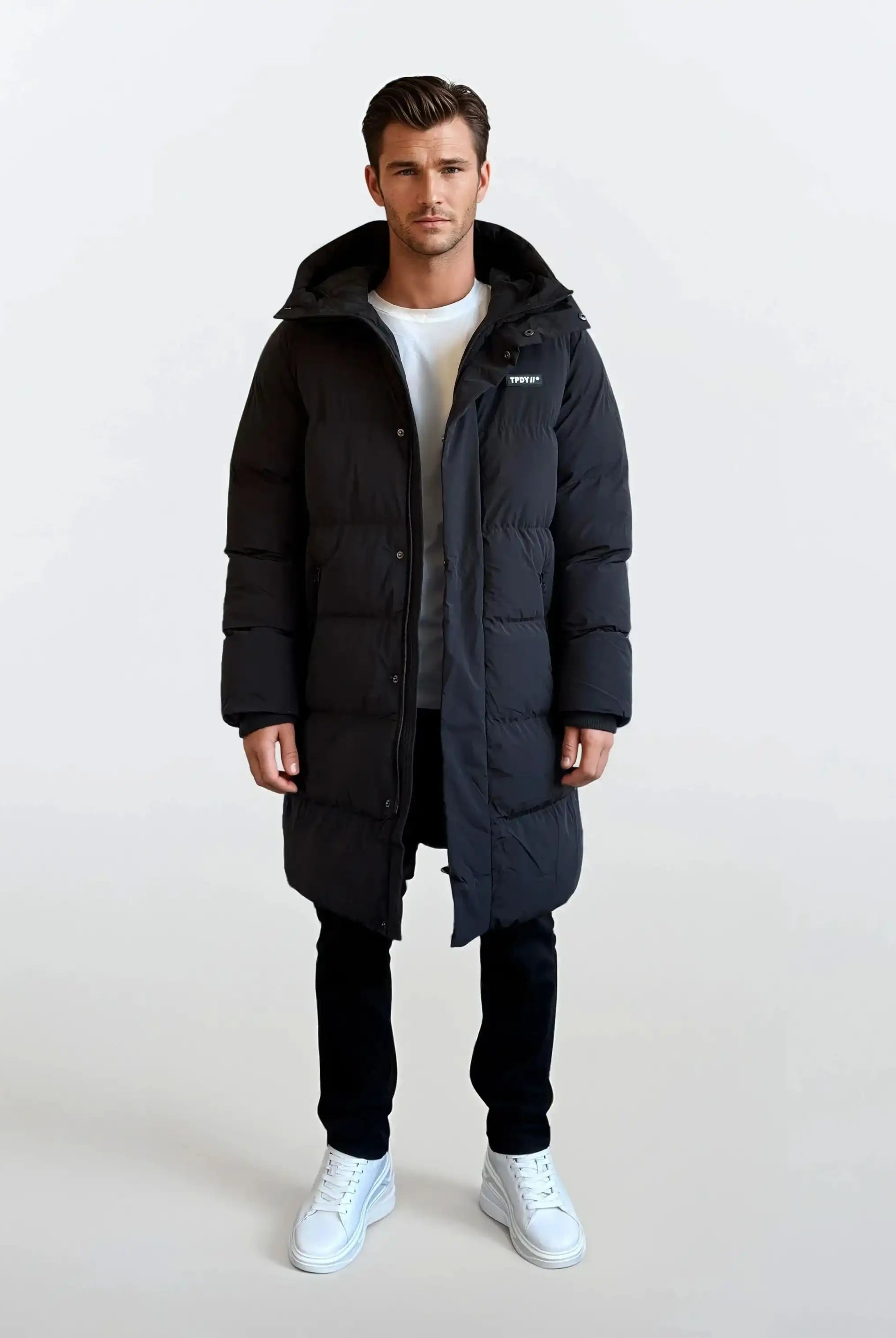 Ein Mann sieht nach vorne und trägt einen trueprodigy Herren Parka mit fester Kapuze im Regular Fit - lang, schwarz und wasserabweisend - über einem weißen T-Shirt, einer schwarzen Hose und weißen Turnschuhen. Der Hintergrund ist einfarbig weiß.